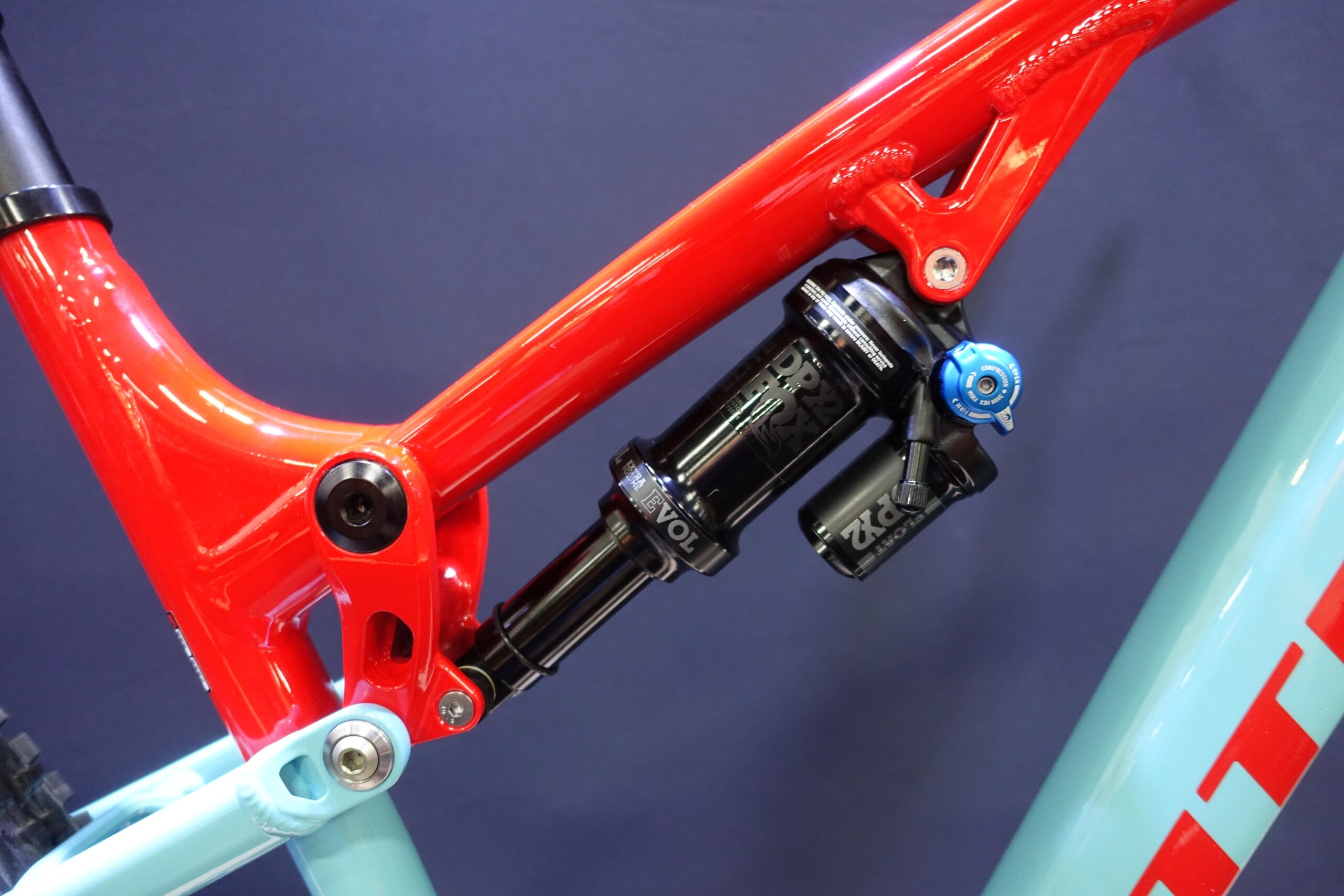Patrol 591: Close-up of a mountain bike frame showcasing a vibrant red section and a black shock absorber with blue adjustment knob, mounted near the back triangle, against a dark background.