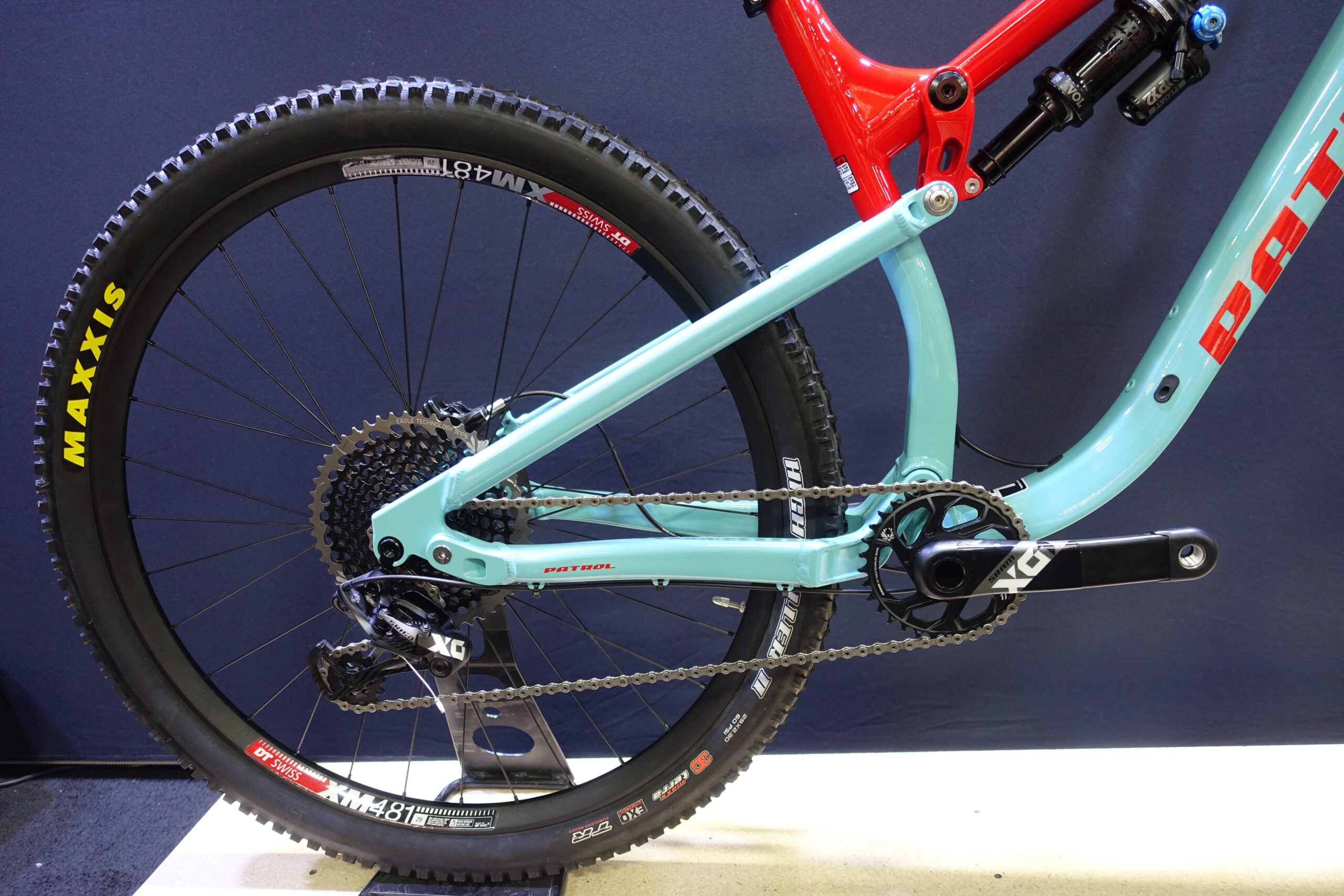 Patrol 591: A close-up view of a mountain bike's rear drivetrain, showing the turquoise frame, red accents, and the Maxxis tire on the wheel. The image includes details of the chain, cassette, and crankset, highlighting the bike's components. The background is a dark color, providing contrast to the bike's vibrant colors.