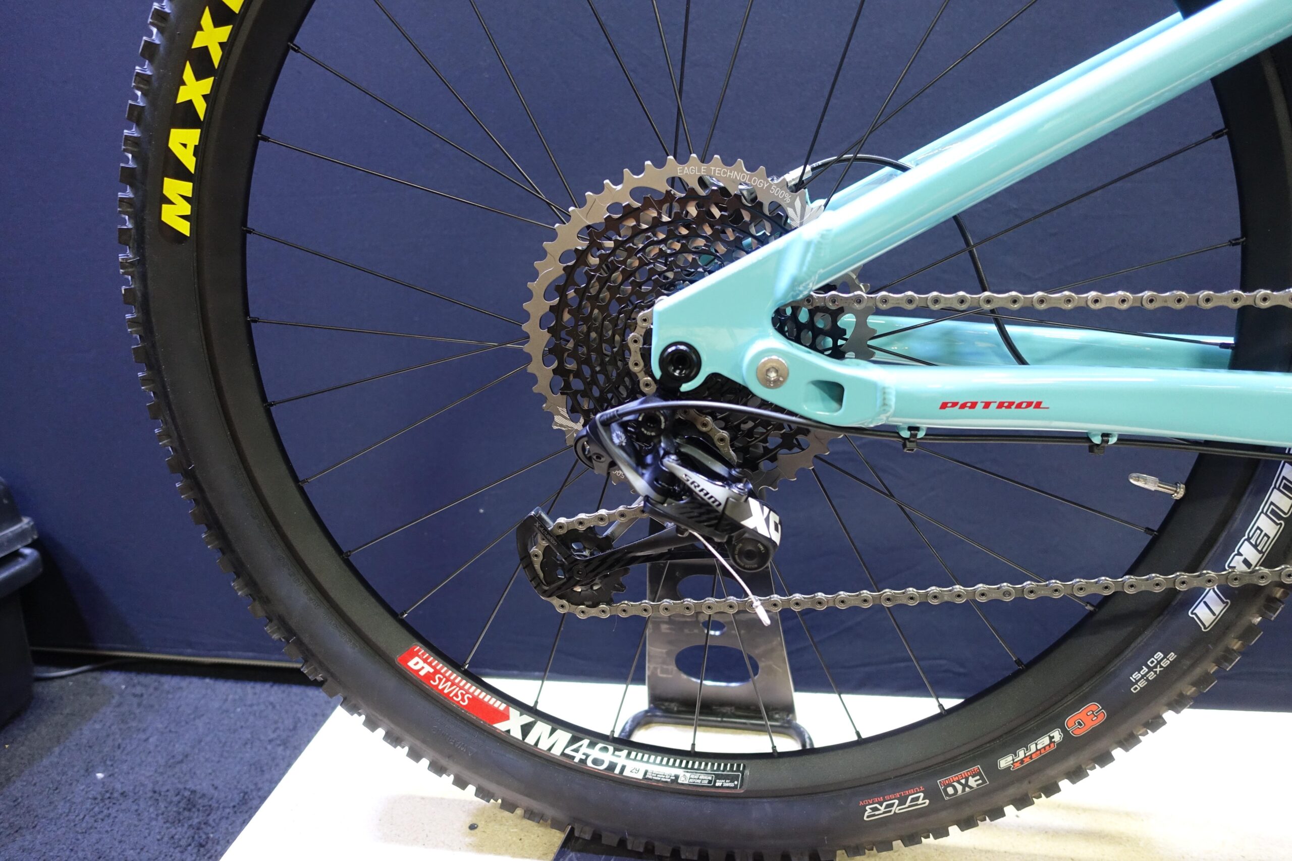 Patrol 591: Close-up image of a mountain bike's rear wheel showing the drivetrain components, including a blue frame, cassette, and derailleur. The tire features the "MAXXIS" logo, and a metal stand supports the bike. The background is a simple dark color to highlight the bike's features.