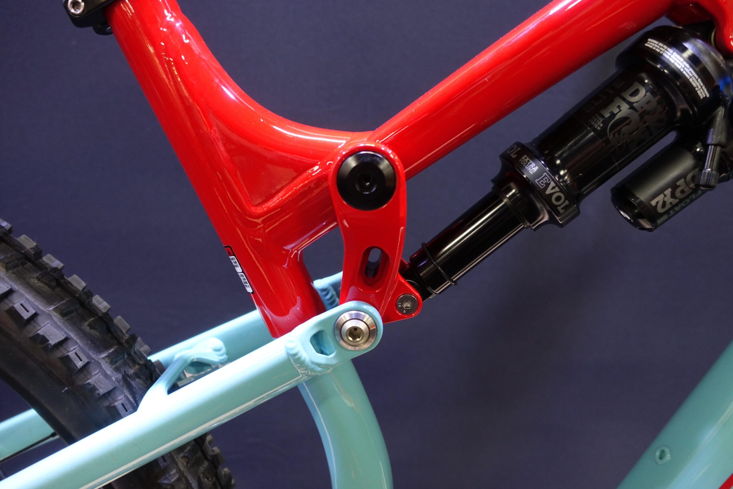 Patrol 591: Close-up view of a bicycle frame featuring a vibrant red upper section and a light blue lower section. The frame displays intricate design details, including a black rear shock absorber. The tire tread is partially visible in the background, emphasizing the bike's rugged design.