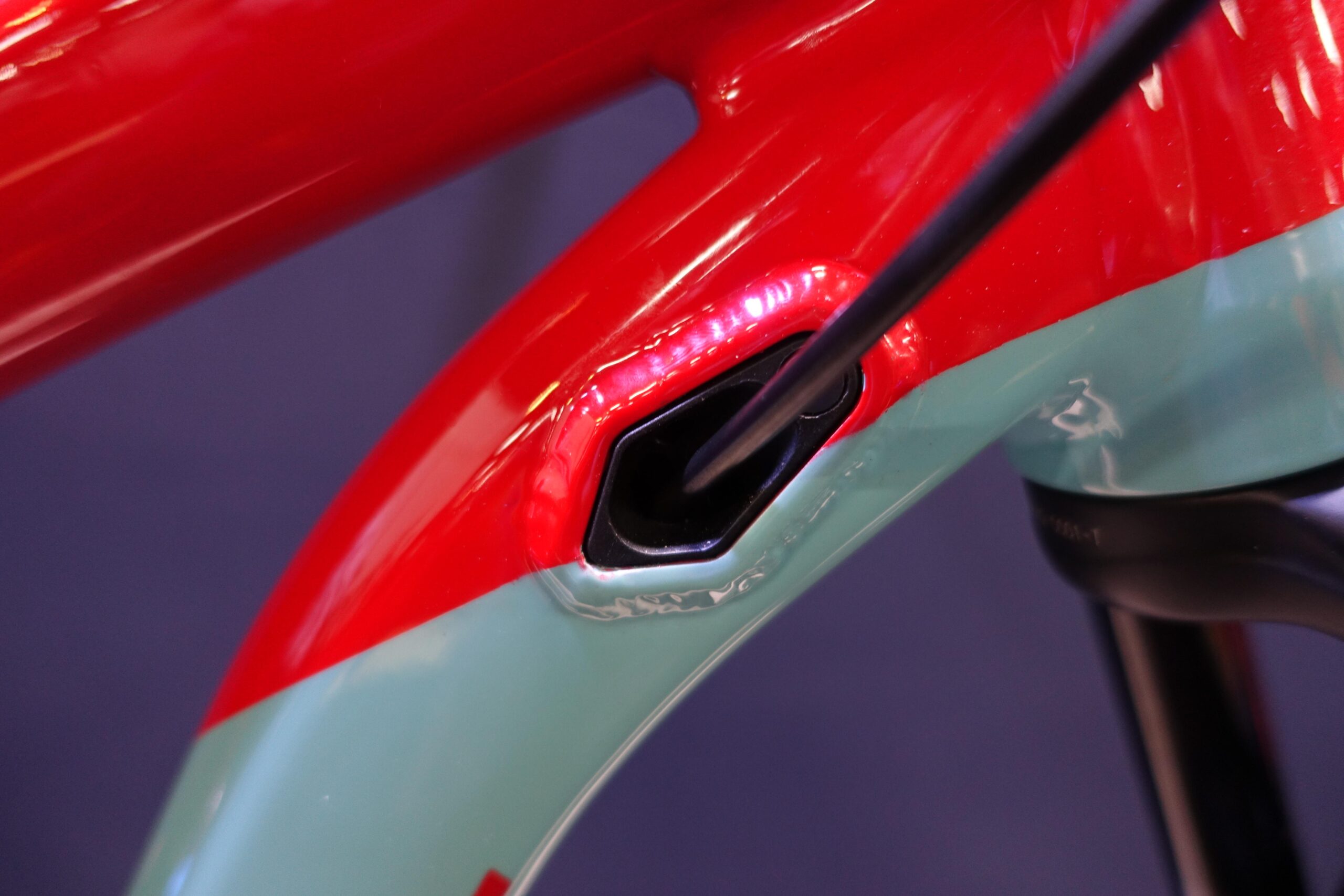 Patrol 591: Close-up view of a bike frame featuring a mix of red and teal paint. The image highlights a cable entry point on the frame, showing details of the smooth finish and design.