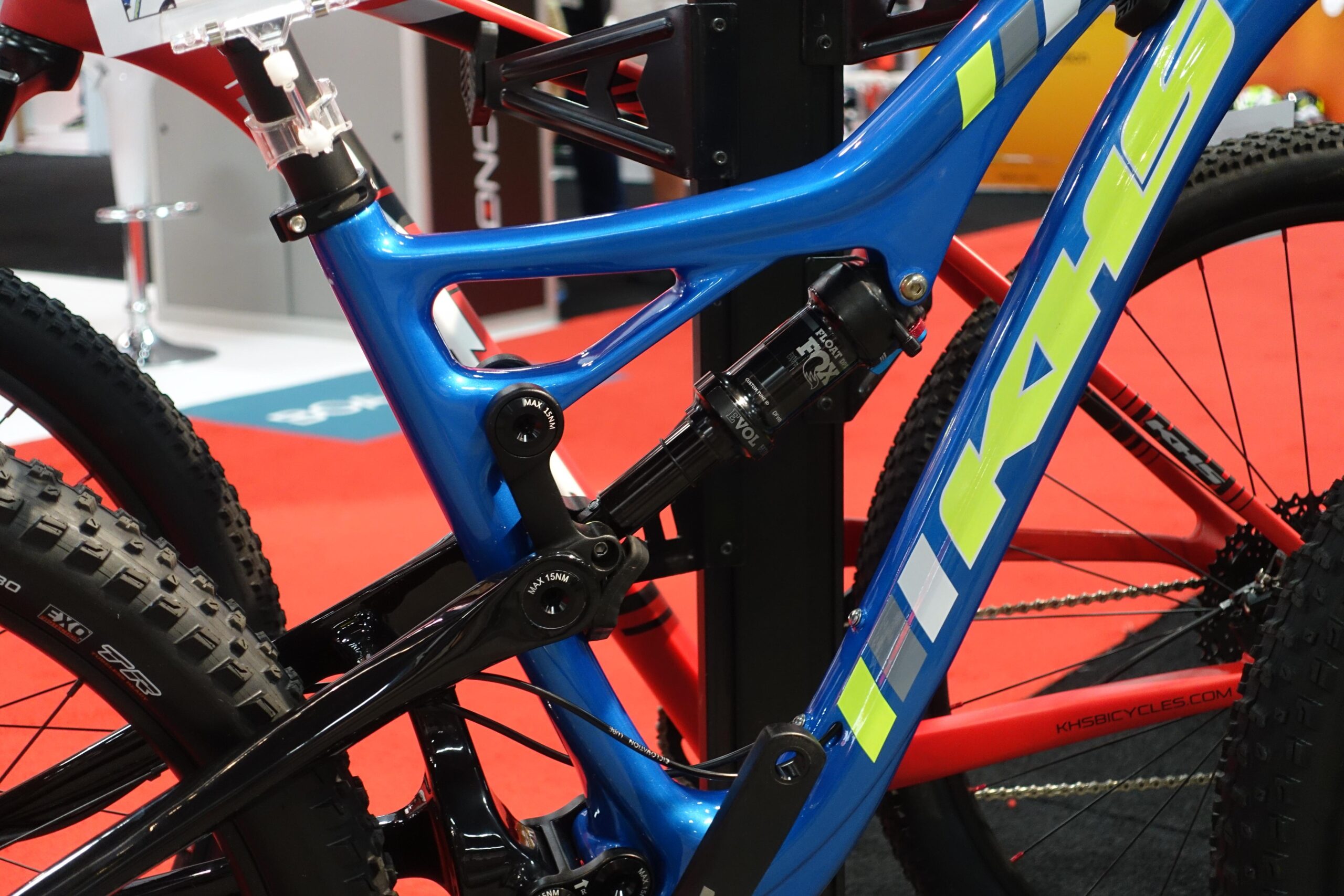 KHS sixfifty 6600: Close-up of a high-performance mountain bike with a blue and black frame. The bike features a dual-suspension system, highlighted by a prominent shock absorber. The background includes a red exhibition floor, suggesting it’s displayed at an event or trade show.
