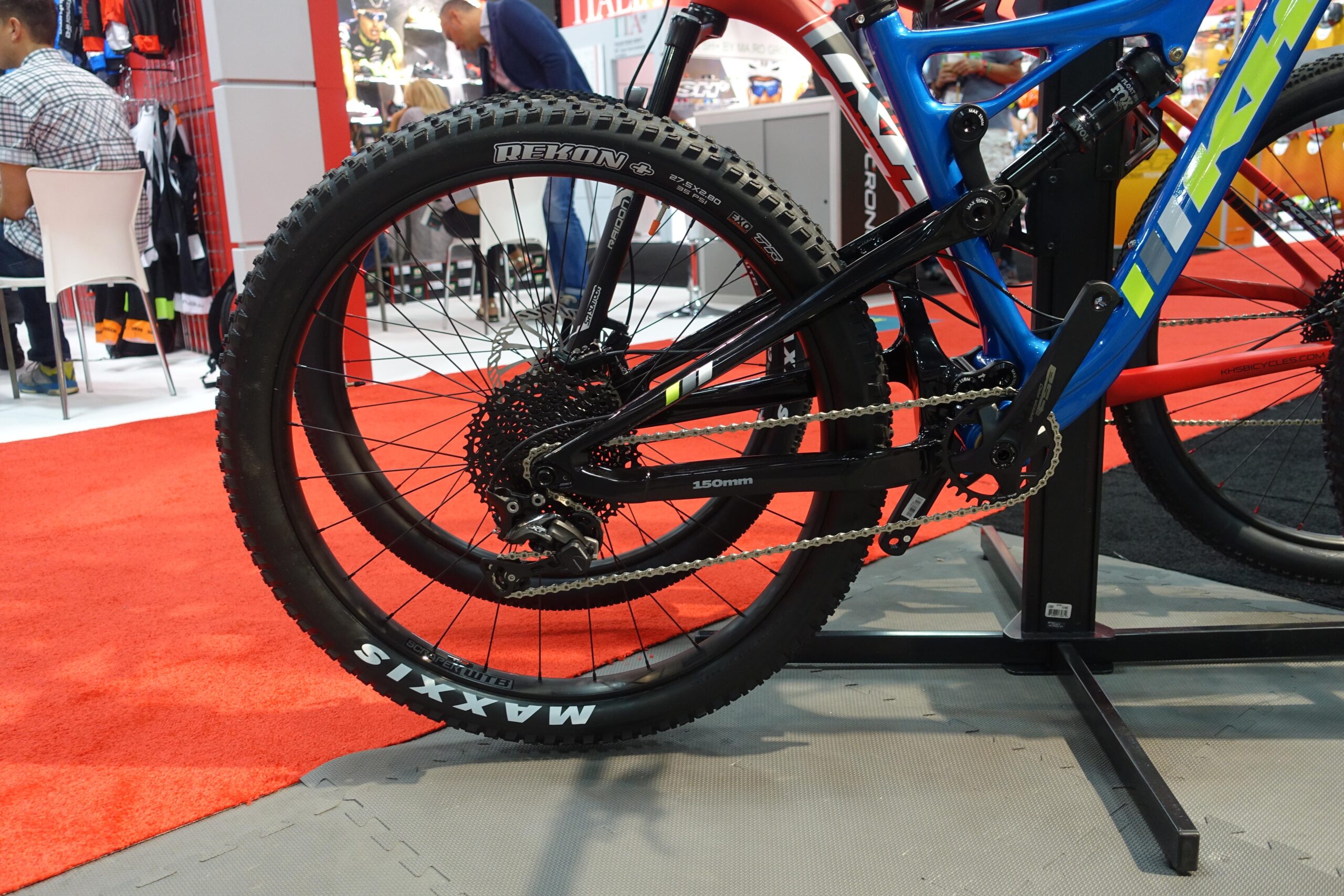 KHS sixfifty 6600: A close-up view of the rear wheel and drivetrain of a mountain bike displayed at an exhibition. The bike features a blue and black frame with colorful accents, and the wheel is equipped with a Maxxis Rekon tire. The background shows blurred elements of a trade show, including people and various bike accessories on display. The floor is covered with red carpet and gray matting.