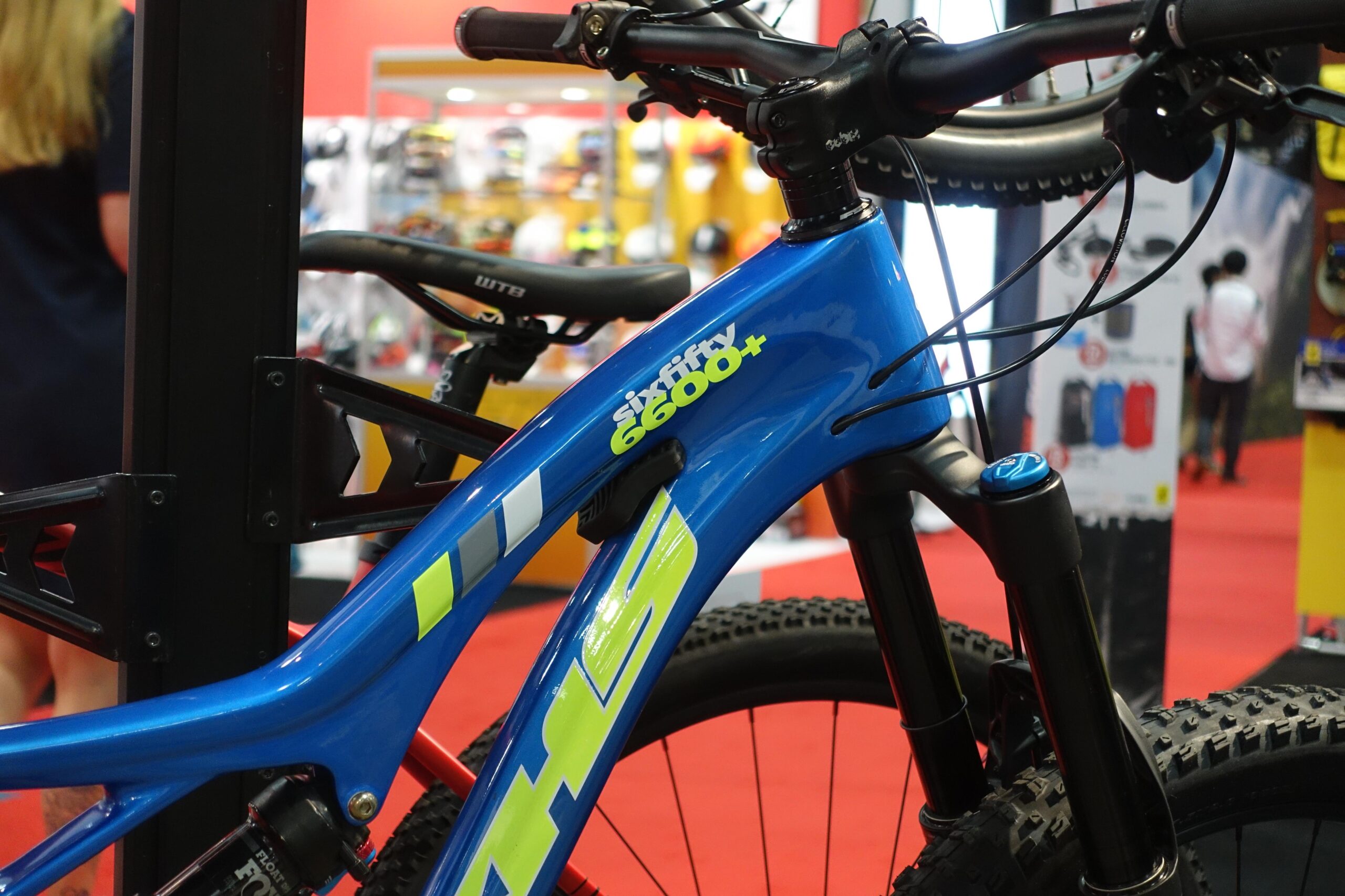 KHS sixfifty 6600: A close-up view of a blue mountain bike featuring the model name "six fifty 6600+" printed in yellow on the frame. The handlebars and front suspension are also visible, along with a blurred background displaying other biking gear and accessories in a trade show setting.