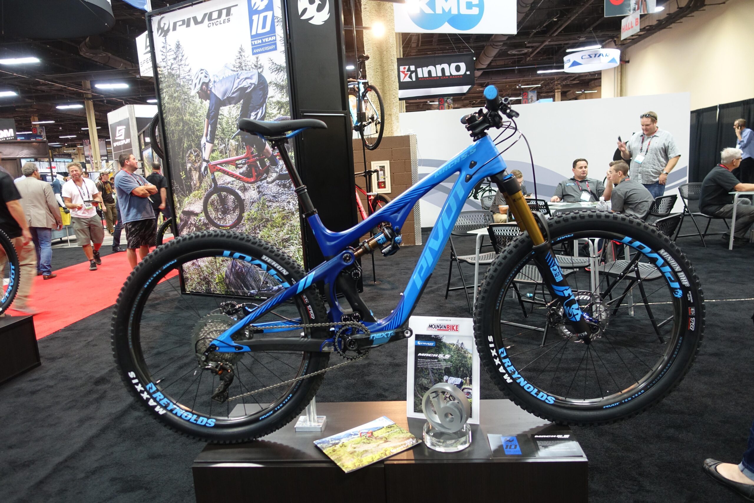 Pivot Mach 5.5: A bright blue mountain bike displayed on a stand at a trade show, featuring the brand name "Pivot" prominently on the frame. The bike has large tires and a suspension system. In the background, attendees can be seen interacting, with various banners and displays related to cycling equipment.