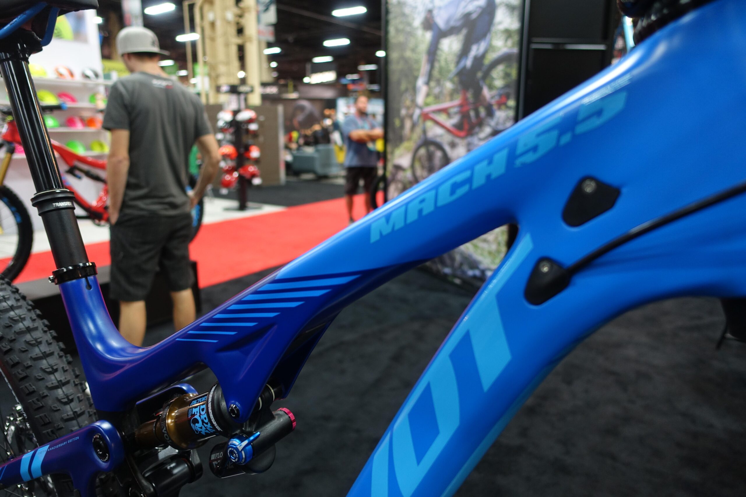 Pivot Mach 5.5: Close-up view of a blue mountain bike frame displaying the model name "Mach 5.5," with visible shock components. A blurred figure of a person in casual attire is seen in the background, alongside colorful bicycles and a trade show environment.