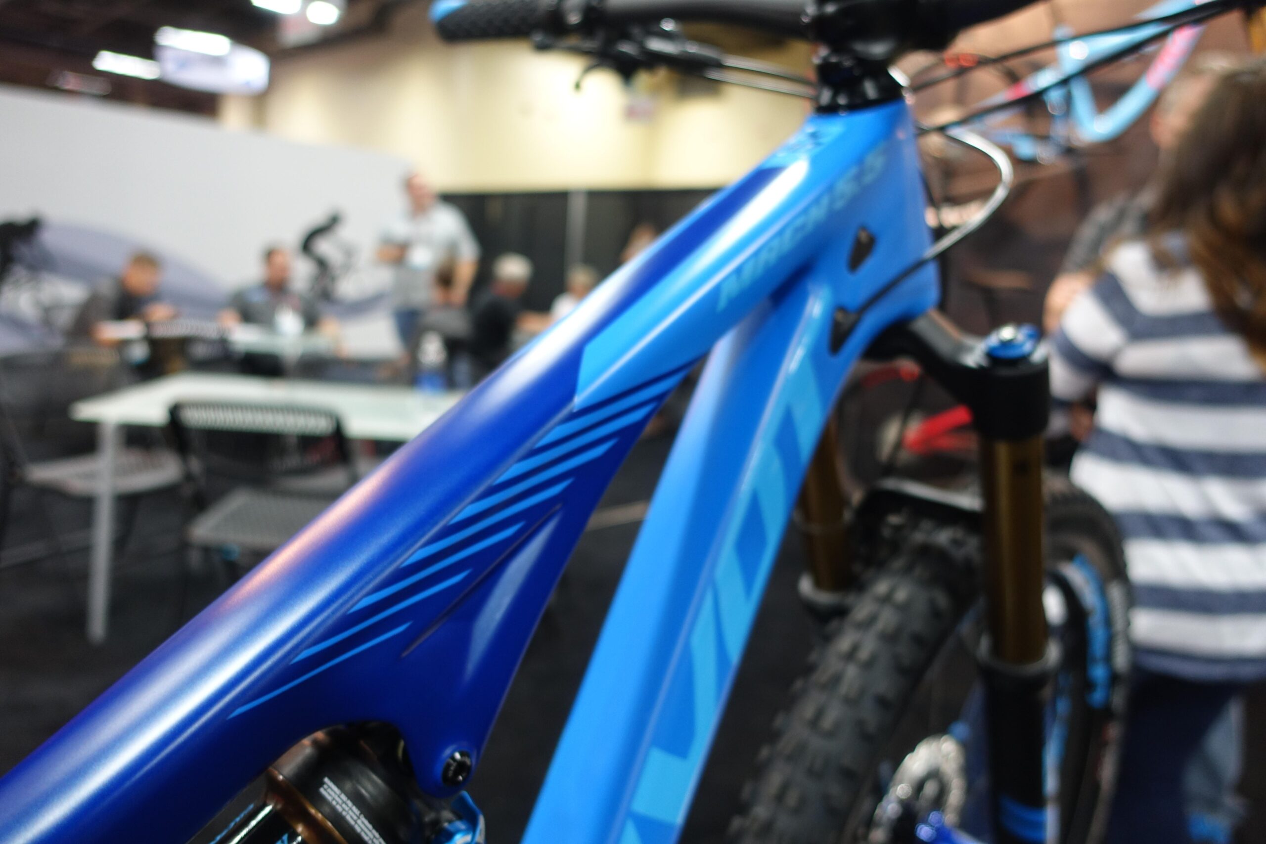 Pivot Mach 5.5: Close-up view of a blue bicycle frame with dynamic graphic design elements. In the background, a blurred group of people are seated at tables, indicating a trade show or exhibition setting.