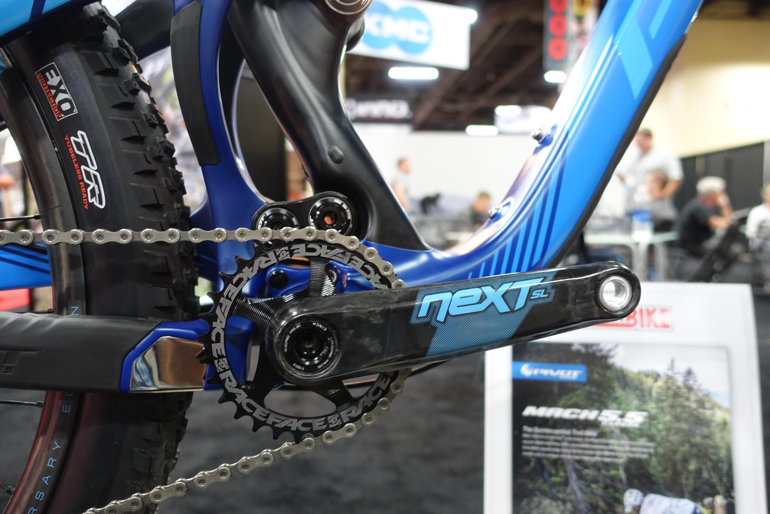 Pivot Mach 5.5: Close-up view of a mountain bike's crankset and chain, showcasing a blue and black frame design with the "next SL" logo on the crank arm. The image also features a detailed view of the chainring, chain, and a specialized tire in the background, set against a blurred trade show environment.