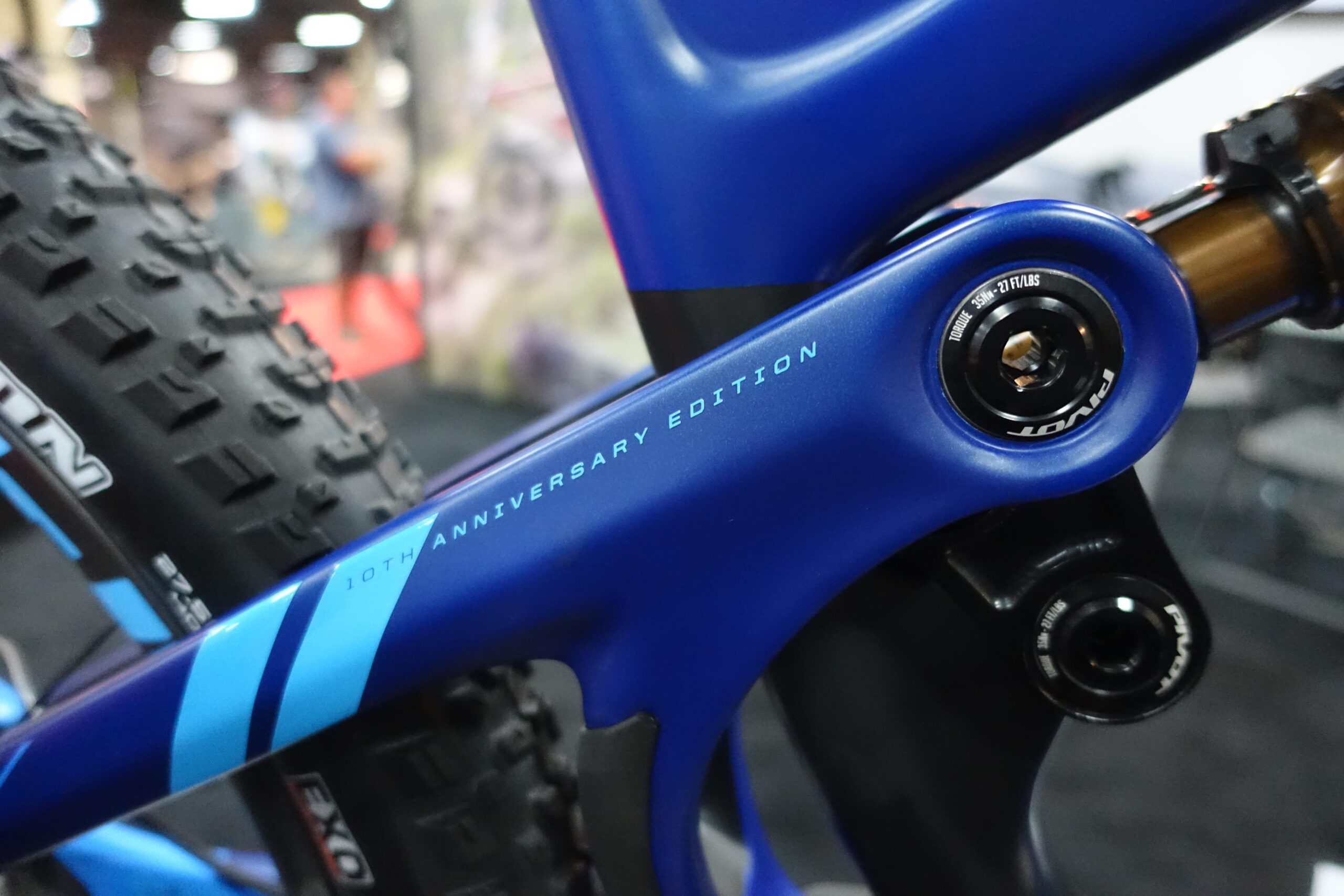 Pivot Mach 5.5: Close-up of a blue bicycle frame featuring a "10th Anniversary Edition" label. The image highlights a section of the frame with a shock absorber and a tire with a rugged tread pattern in the background, set in an indoor environment.