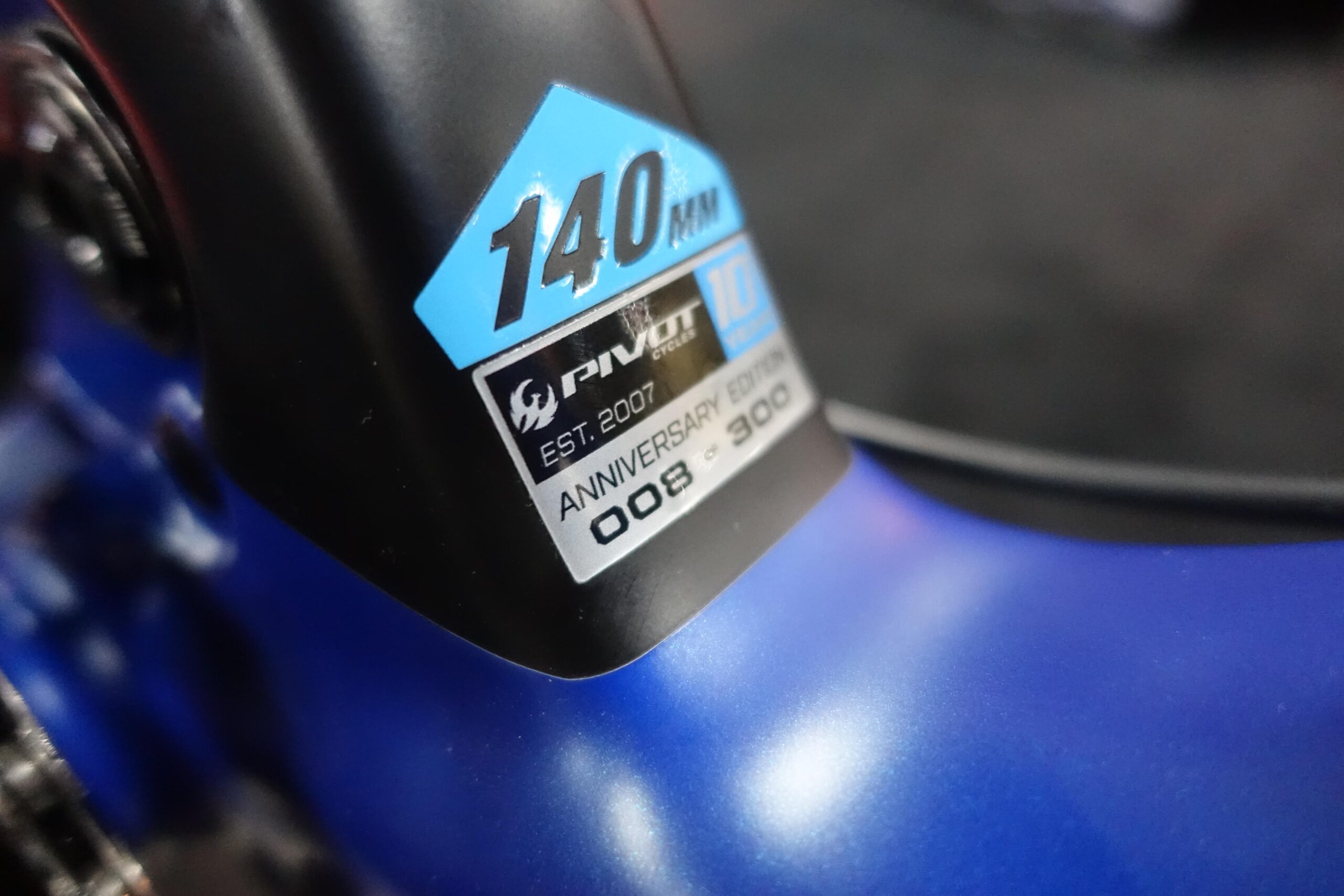 Pivot Mach 5.5: Close-up of a bicycle frame showcasing a blue and black sticker that reads "140mm" and "Pivot Cycles." The sticker notes "EST. 2007" and indicates it is an "Anniversary Edition" with a limited number designation of "008 - 300." The background is softly blurred, highlighting the details of the sticker.