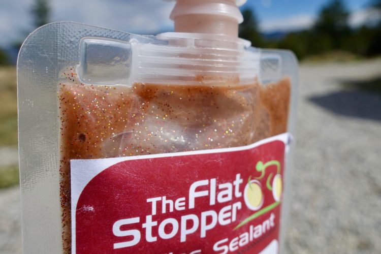 Close-up of a pouch of tire sealant with a red label reading "The Flat Stopper." The pouch contains a glittery, brown liquid and is partially transparent, revealing the contents inside. The background shows a blurred outdoor setting with trees and a blue sky.