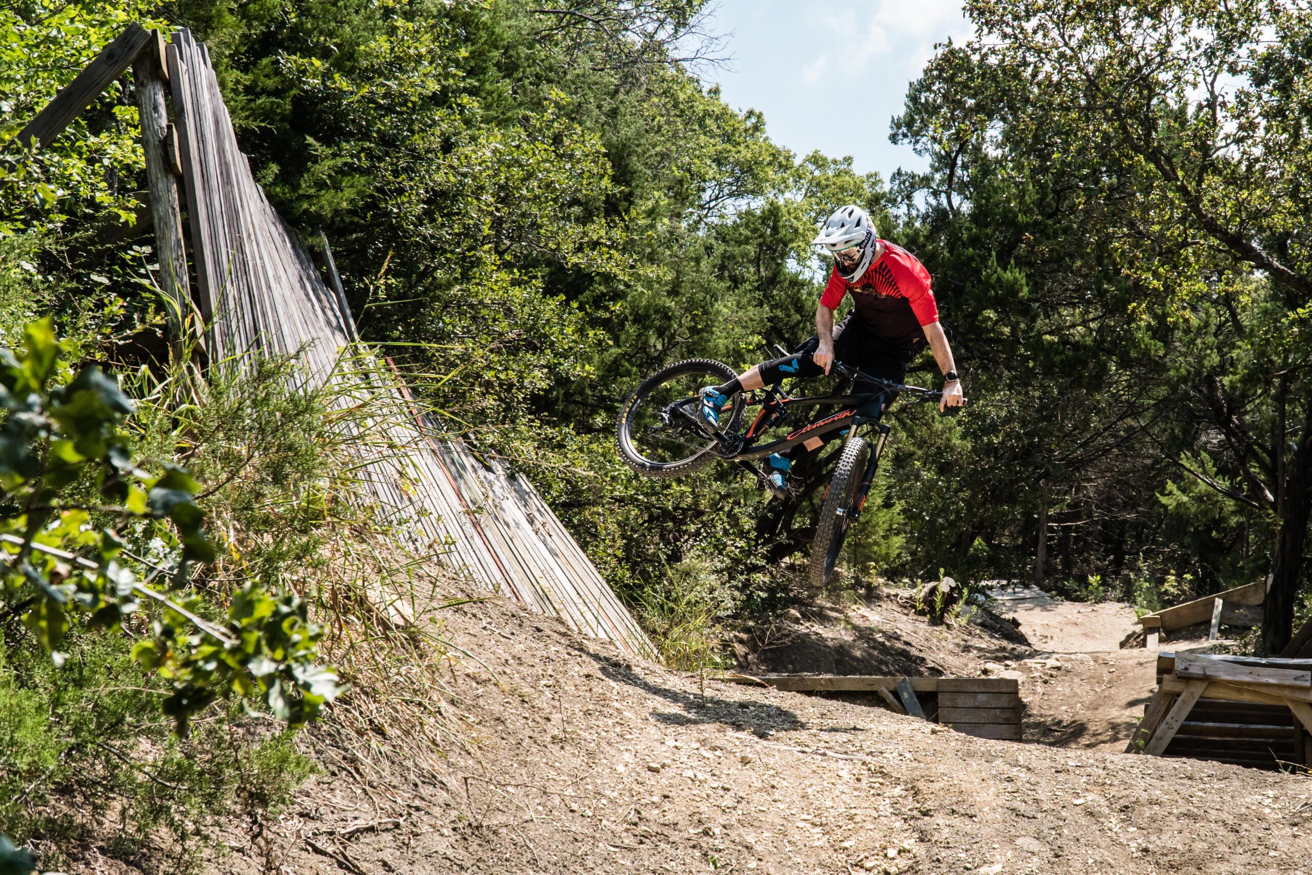 Ellsworth Rogue: A mountain biker performing a jump over a wooden ramp on a natural dirt trail, surrounded by greenery and trees. The rider is wearing a red jersey and protective gear, showcasing dynamic action in a bright, sunny environment.