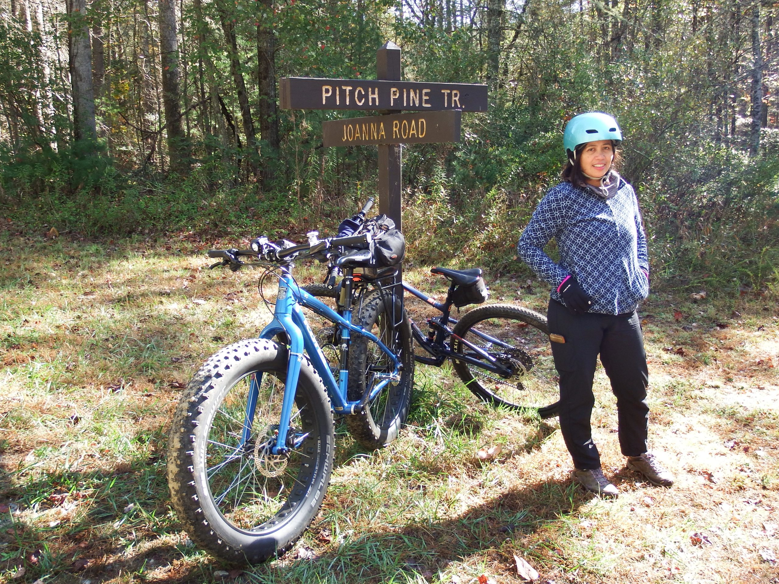 A person wearing a helmet and casual outdoor clothing stands near two parked bicycles beside a trail sign that reads "Pitch Pine Tr. Joanna Road." The setting features a sunny day with a grassy area surrounded by trees. DuPont State Recreational Forest mountain bike trail.