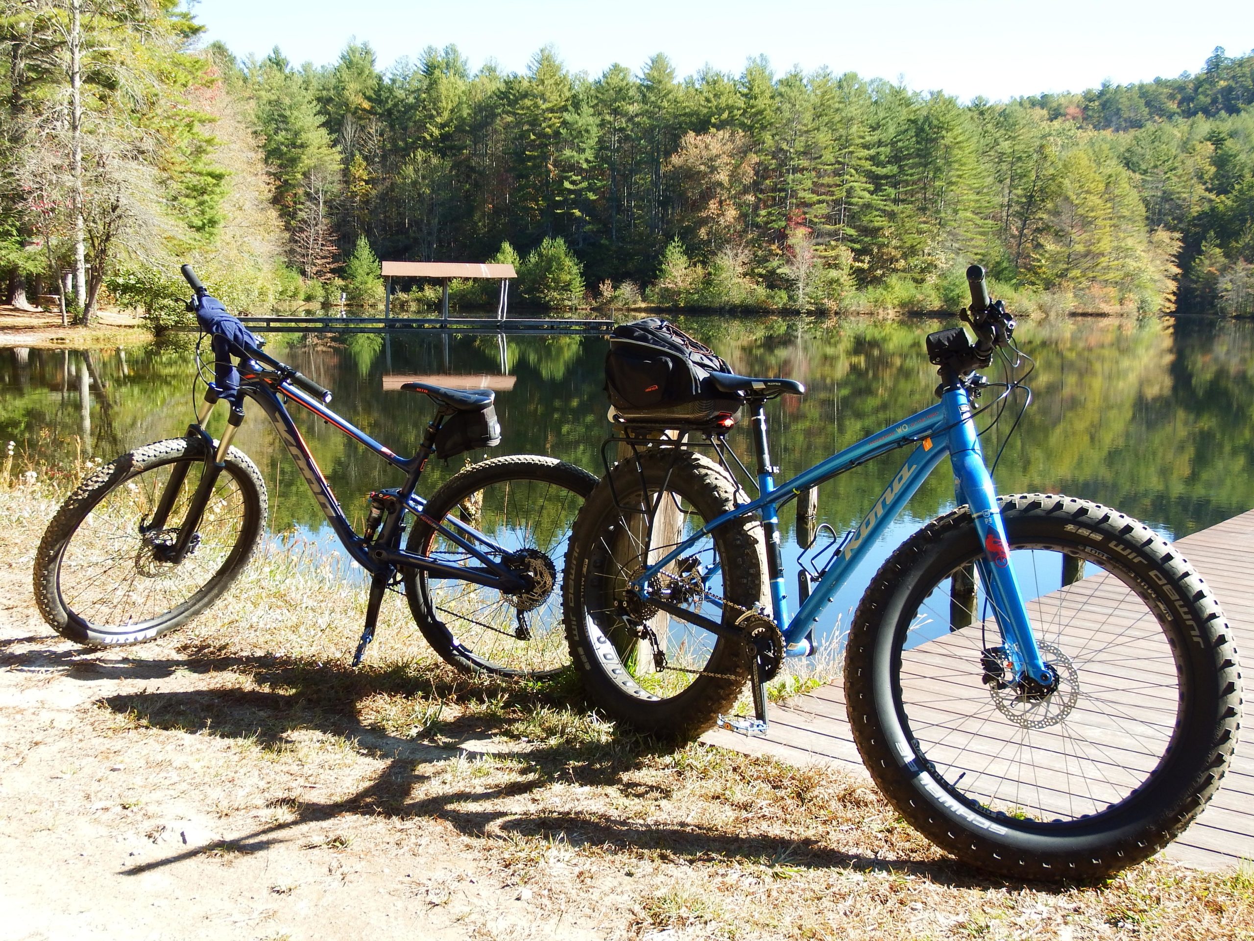 Two mountain bikes are parked next to a calm lake surrounded by trees. The bike on the left is a dark color with a sleek design, while the one on the right is bright blue with thick tires. In the background, a wooden dock extends over the water, and a small shelter can be seen nestled among the trees. The scene is illuminated by bright sunlight, showcasing a peaceful outdoor setting. DuPont State Forest mountain bike trail.