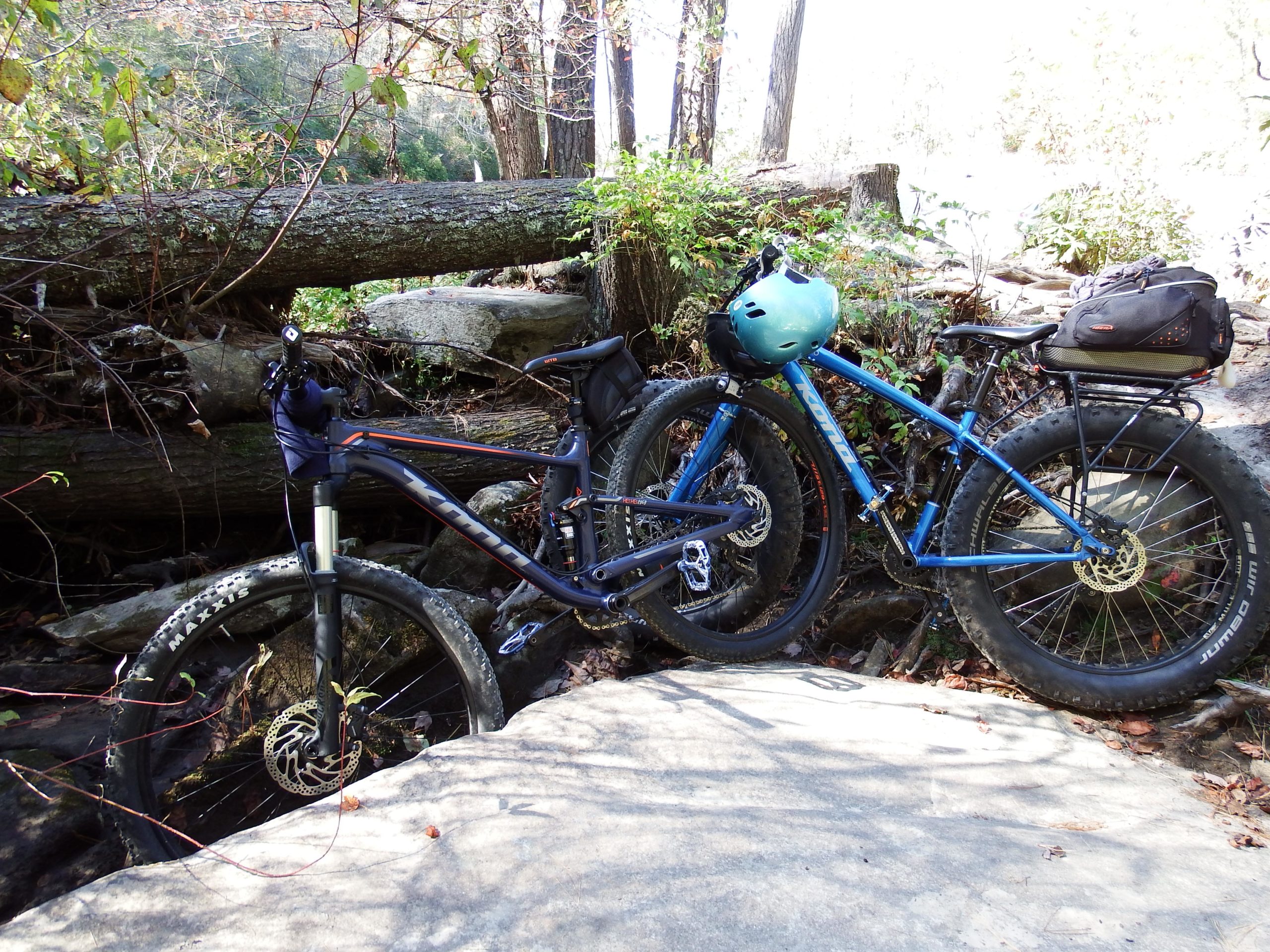 Two mountain bikes are parked alongside a rocky path in a forested area. One bike is dark with bright orange accents, while the other is blue with a light blue helmet attached. Surrounding the bikes are fallen logs, greenery, and rocky terrain, indicating a natural outdoor setting perfect for biking adventures. DuPont State Forest mountain bike trail.