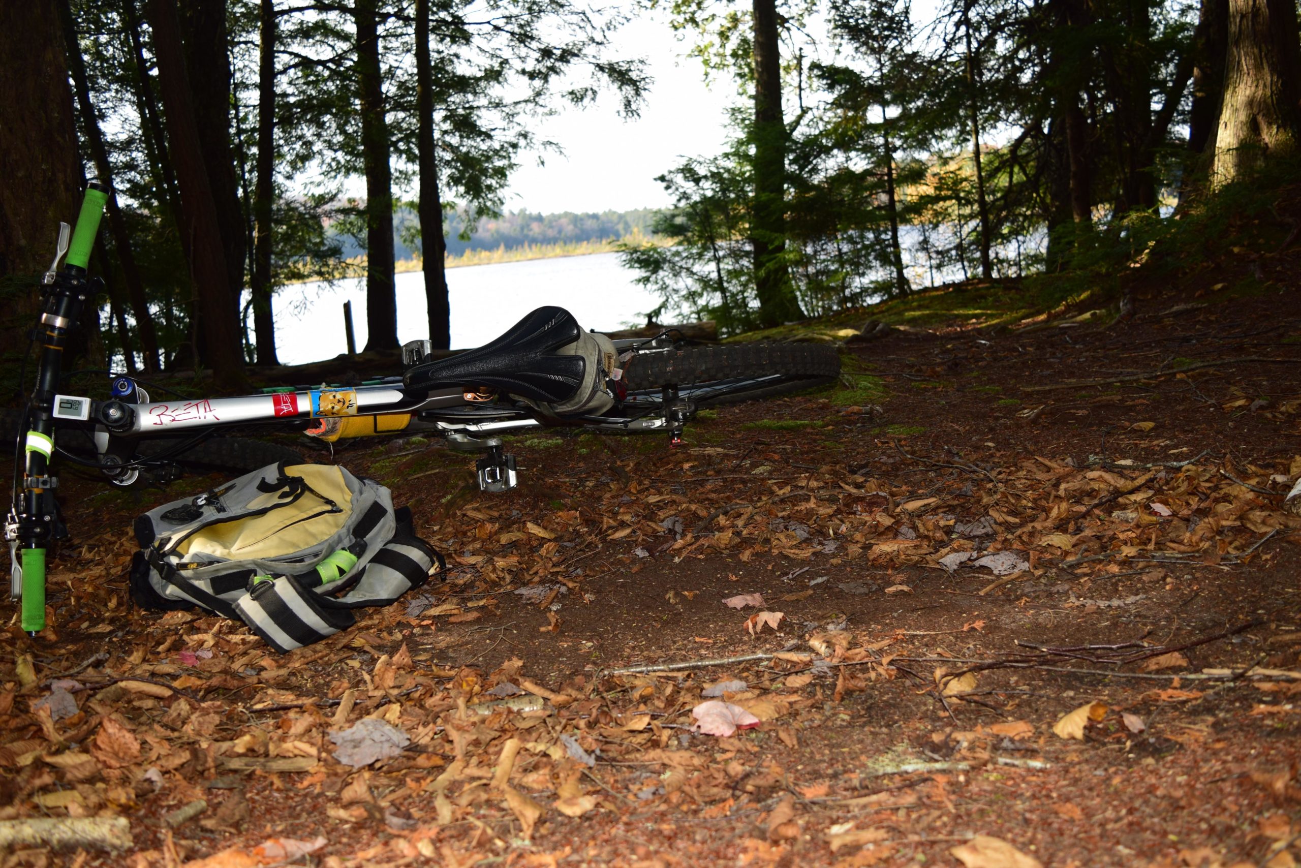 A mountain bike and a backpack resting on the forest floor, surrounded by fallen leaves. In the background, a tranquil lake can be seen peeking through the trees, with hints of autumn colors in the foliage. The scene conveys a peaceful outdoor atmosphere, ideal for biking and nature exploration. Lizzie Oliver Trail mountain bike trail.