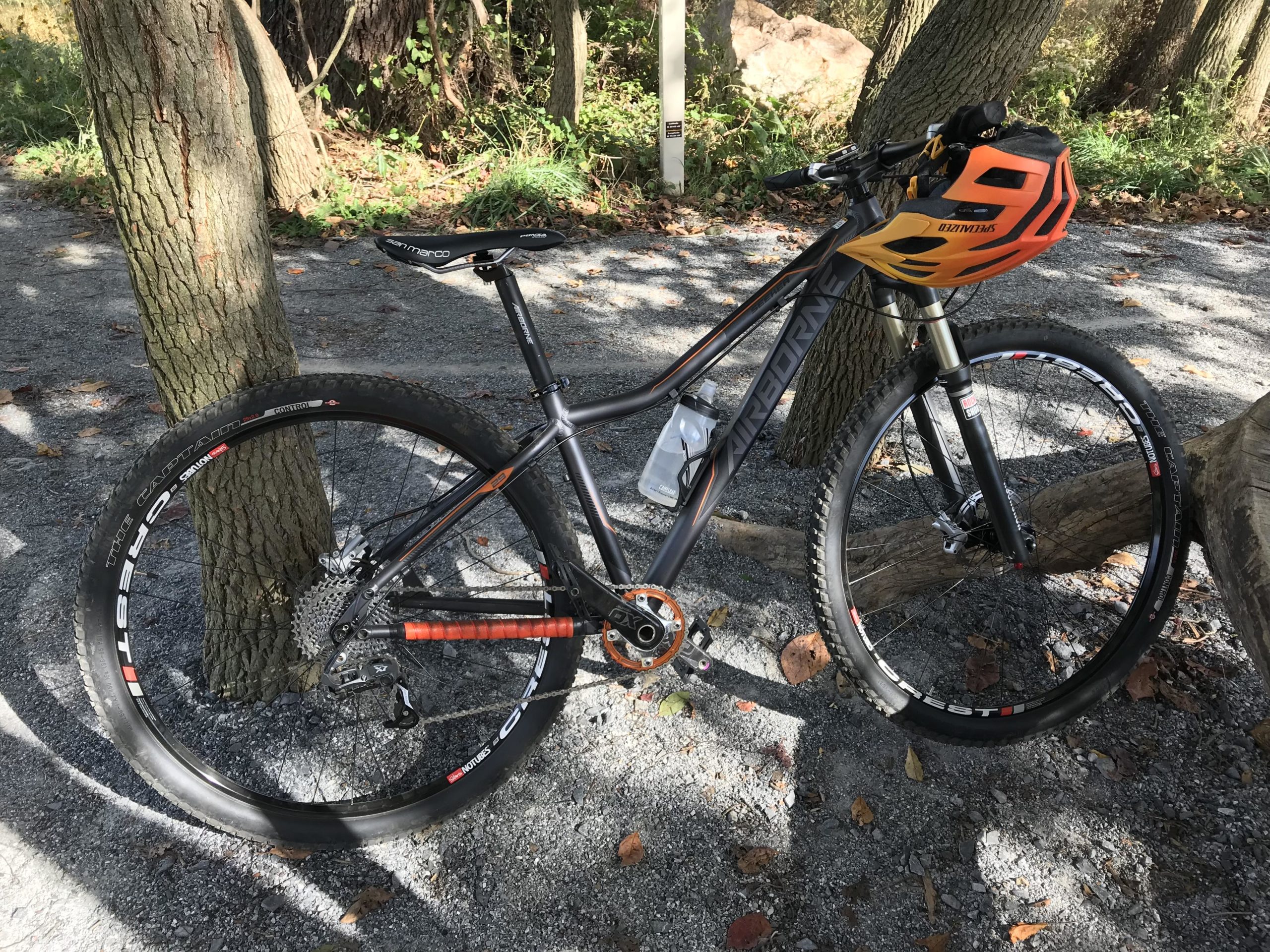A mountain bike resting against a tree on a gravel path, featuring black and orange accents, wide tires, and a matching orange helmet secured on the handlebars. The background includes trees and fallen leaves, suggesting a natural outdoor setting. White Clay Creek mountain bike trail.