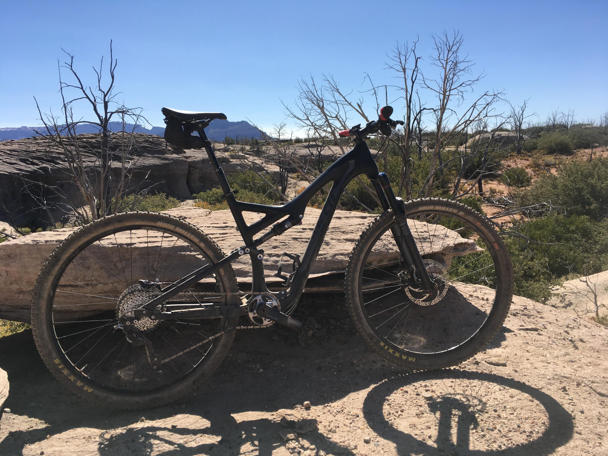 Specialized Stumpjumper FSR comp carbon 650B: A mountain bike resting on a rock formation in a desert landscape, with dry bushes and distant mountains visible under a clear blue sky. The bike is black with visible dirt on the tires, showcasing its off-road use.