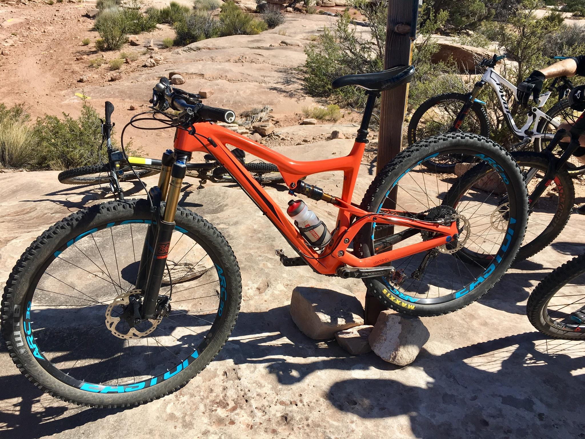 Ibis Ripley: An orange mountain bike with black and blue accents, resting on rocky terrain with a water bottle attached to the frame. In the background, another bike is partially visible among desert vegetation.