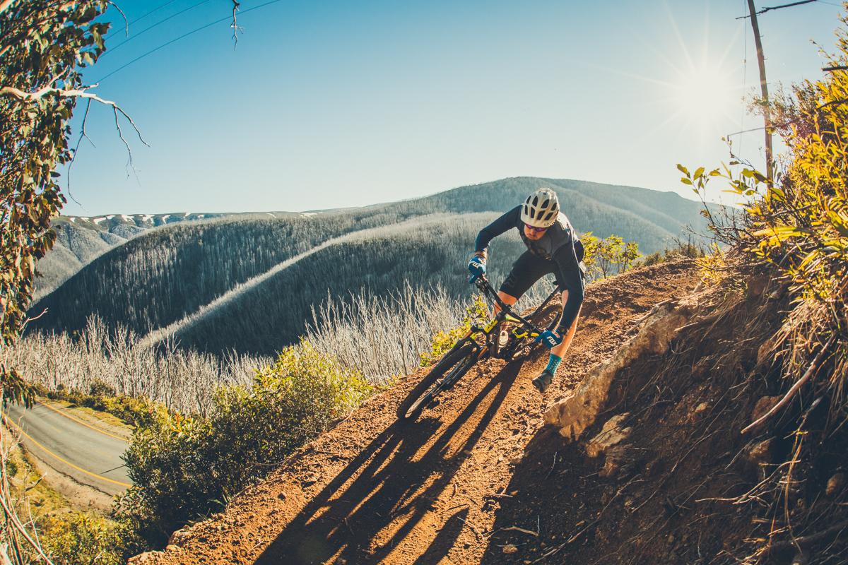 A mountain biker navigating a winding dirt trail on a scenic hillside, with mountains in the background and the sun shining brightly in the sky. The biker is leaning into the turn, showcasing dynamic movement and the thrill of the ride. Lush greenery and a road can be seen alongside the trail. Flowtown mountain bike trail.