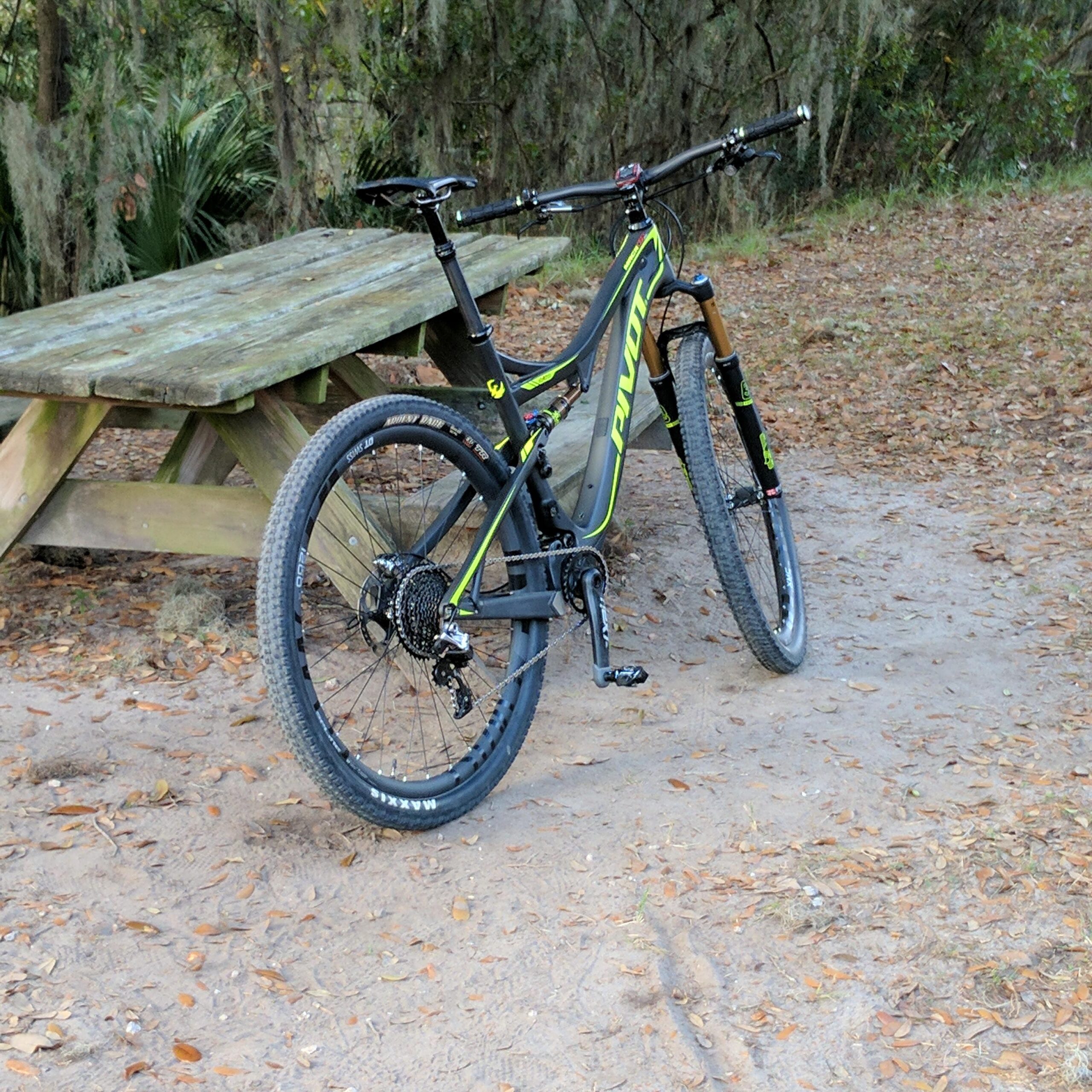 Pivot Mach 429SL Carbon: A modern mountain bike with a black and neon green frame is resting beside a wooden picnic table in a natural setting. The ground is covered in sand and scattered leaves, indicating an outdoor trail environment.