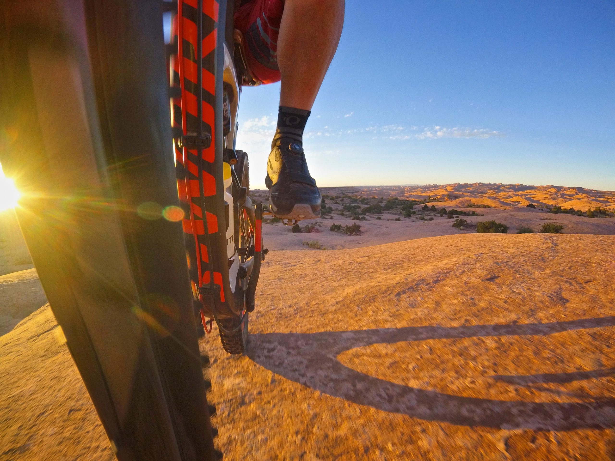 A close-up view of a mountain bike riding over rocky terrain during sunset. The image captures the bike tire, with the sun setting in the background, casting a warm glow and lens flare. The surrounding landscape features rugged hills and open sky. Slickrock mountain bike trail.