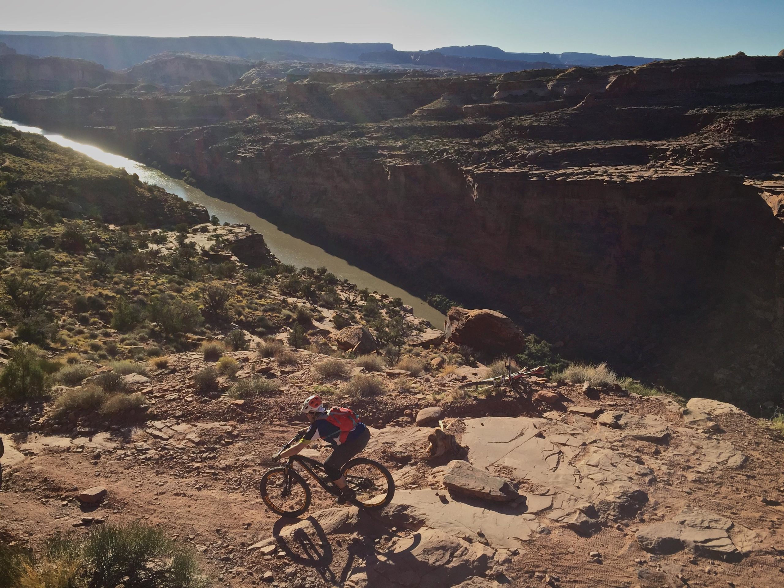 A mountain biker navigates a rocky trail overlooking a winding river in a canyon, surrounded by rugged terrain and greenery. The sun casts a warm glow on the landscape, highlighting the steep cliffs and natural beauty of the area. Porcupine Rim mountain bike trail.
