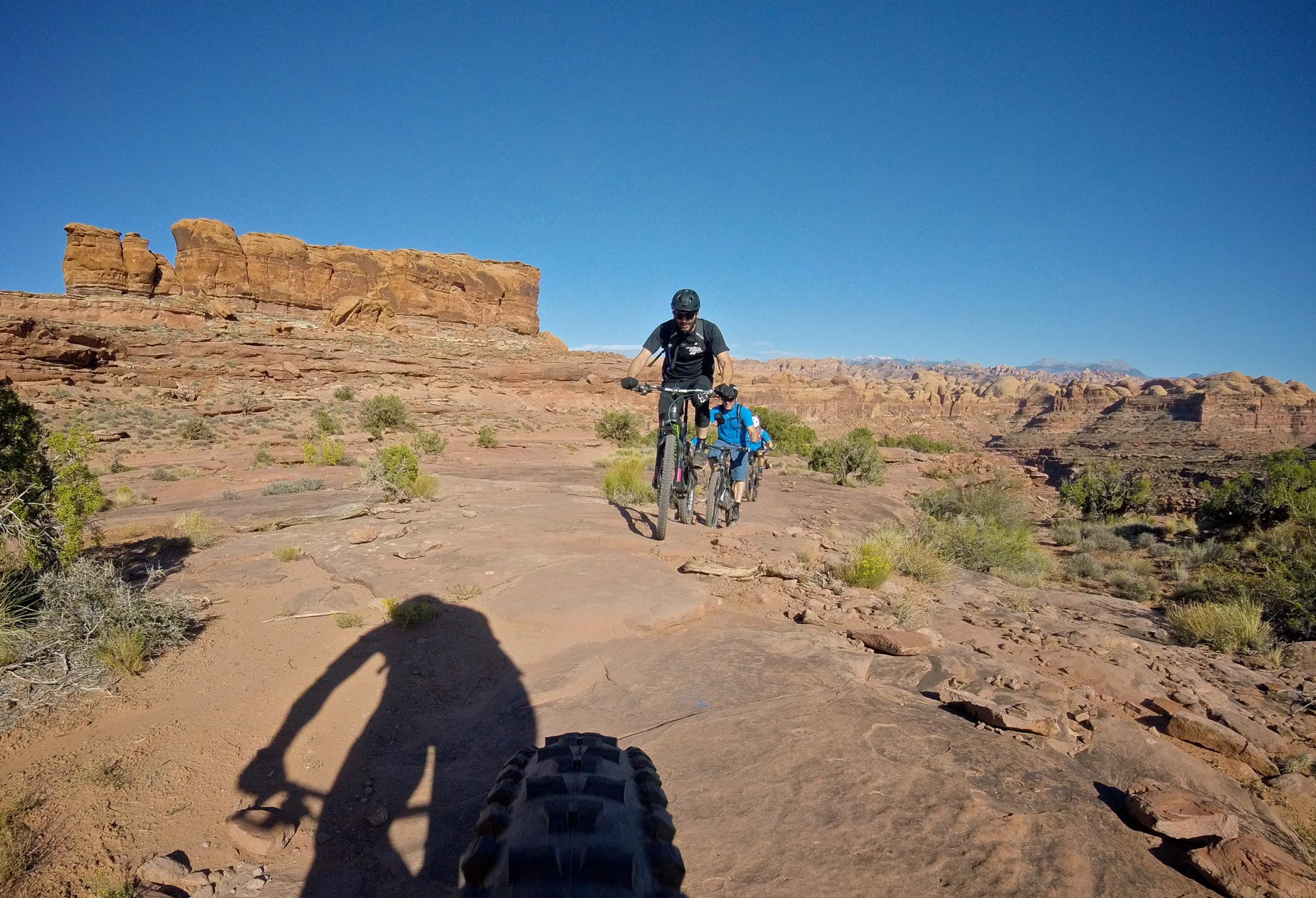 Mountain bikers navigating a rocky trail in a desert landscape, with red rock formations and blue sky in the background. The image captures a sense of adventure and outdoor activity. Hymasa mountain bike trail.