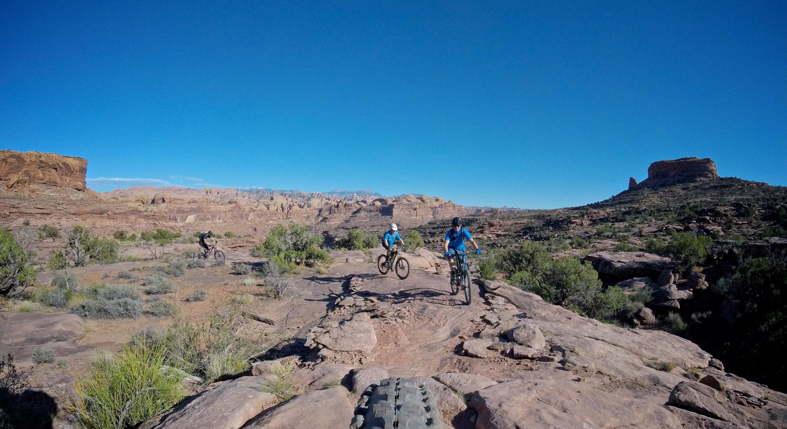 Three mountain bikers riding along a rocky trail in a desert landscape, with vibrant blue skies and colorful rock formations in the background. The trail features sparse vegetation and rugged terrain, capturing the essence of outdoor adventure. Hymasa mountain bike trail.