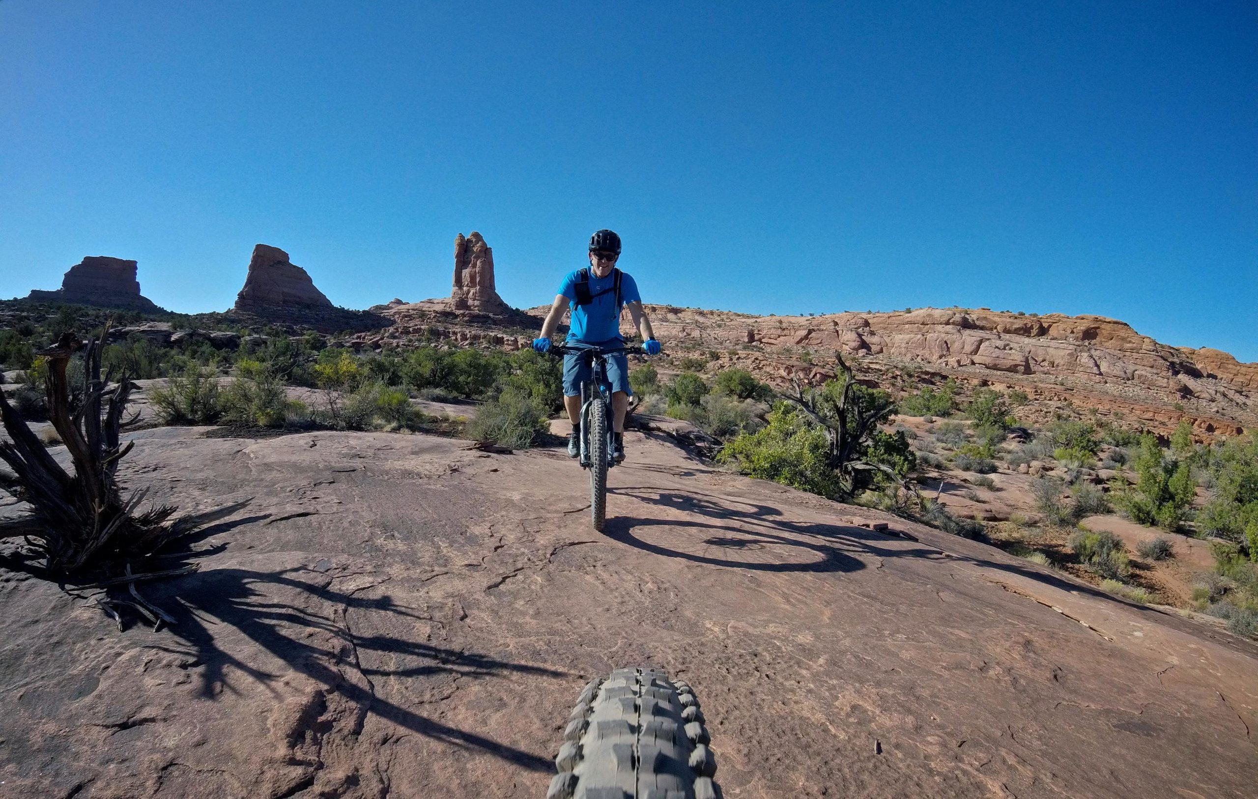 A mountain biker in a blue shirt and helmet rides over rocky terrain, with distinctive rock formations and a clear blue sky in the background. The perspective captures both the rider and the front wheel of the bike. Hymasa mountain bike trail.