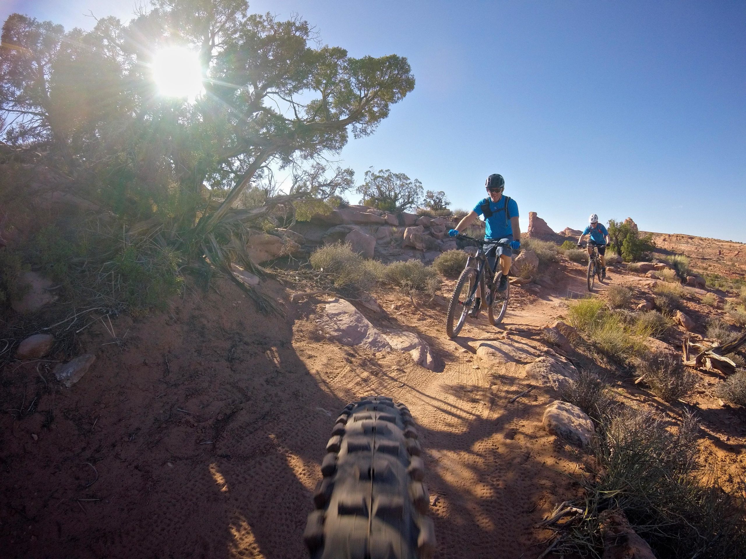 Two mountain bikers navigate a dirt trail in a sunny desert landscape, with a bright sun shining through the branches of a nearby tree. The scene captures the rugged terrain and natural beauty of the surroundings. Hymasa mountain bike trail.