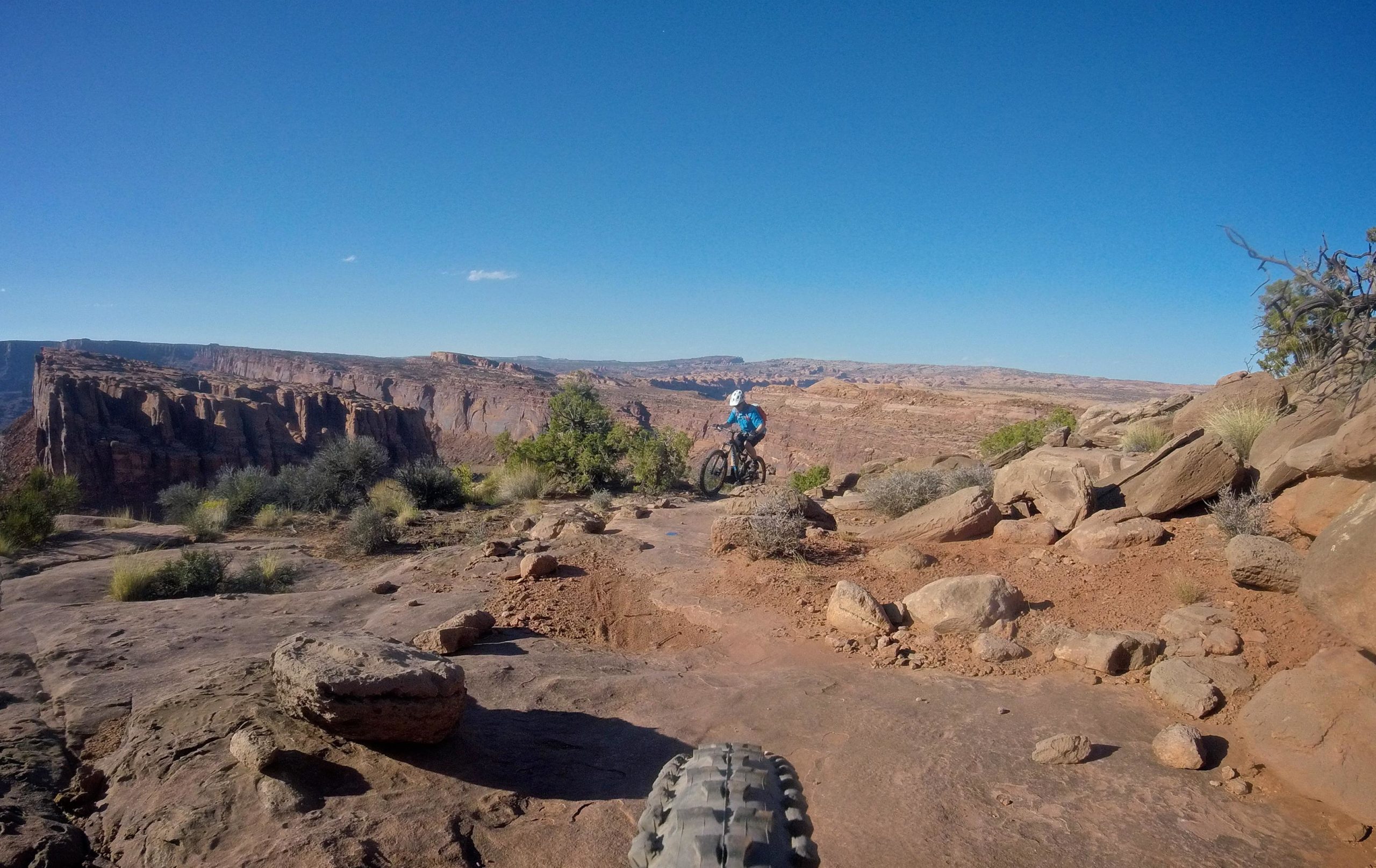 A mountain biker riding on rocky terrain with a panoramic view of cliffs and canyons under a clear blue sky. Captain Ahab mountain bike trail.