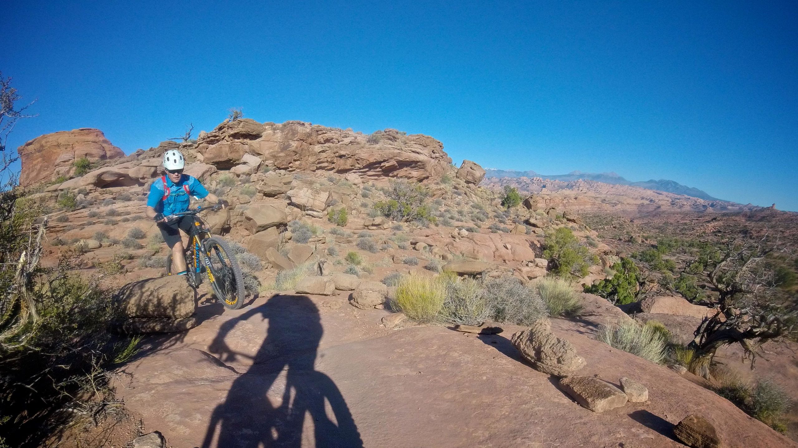 A mountain biker navigates rocky terrain under a clear blue sky, surrounded by desert vegetation and rugged landscapes. The cyclist, wearing a helmet and blue attire, is in motion as he approaches a large rock. Shadows of the bike and rider stretch across the ground. Captain Ahab mountain bike trail.