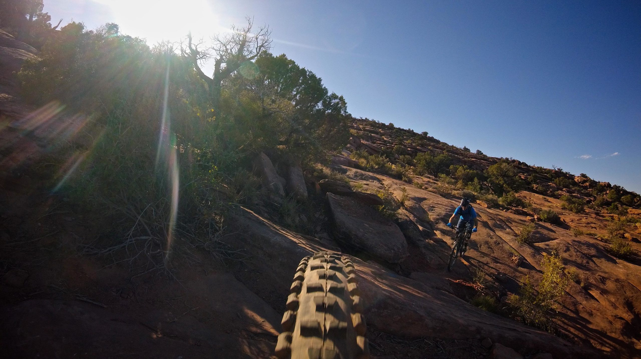 A mountain biker navigating rocky terrain under bright sunlight, with the focus on the bike tire in the foreground and a rider in a blue shirt visible in the background. The landscape features rugged rock formations and sparse vegetation, set against a clear blue sky. Captain Ahab mountain bike trail.