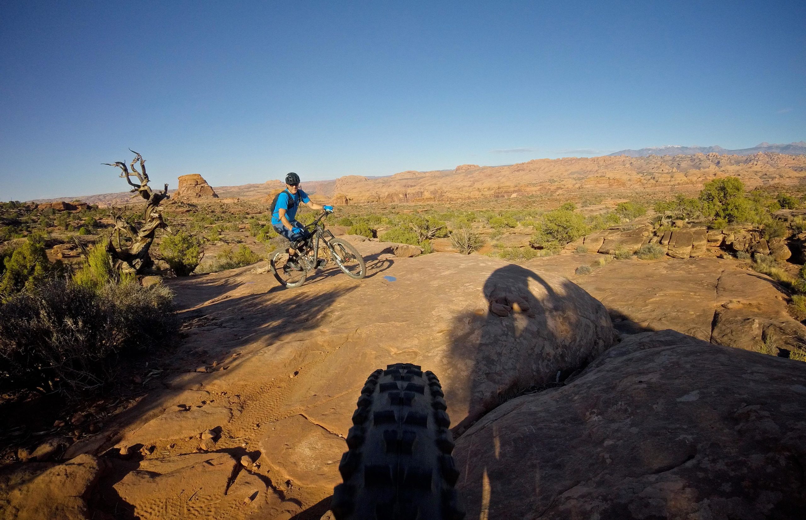 A mountain biker in a blue shirt and helmet rides along a rocky trail in a desert landscape, with red rock formations and sparse vegetation in the background. The foreground shows a close-up of the bike tire on the ground, casting a shadow on the rock surface. The sky is clear and blue, indicating a sunny day. Captain Ahab mountain bike trail.