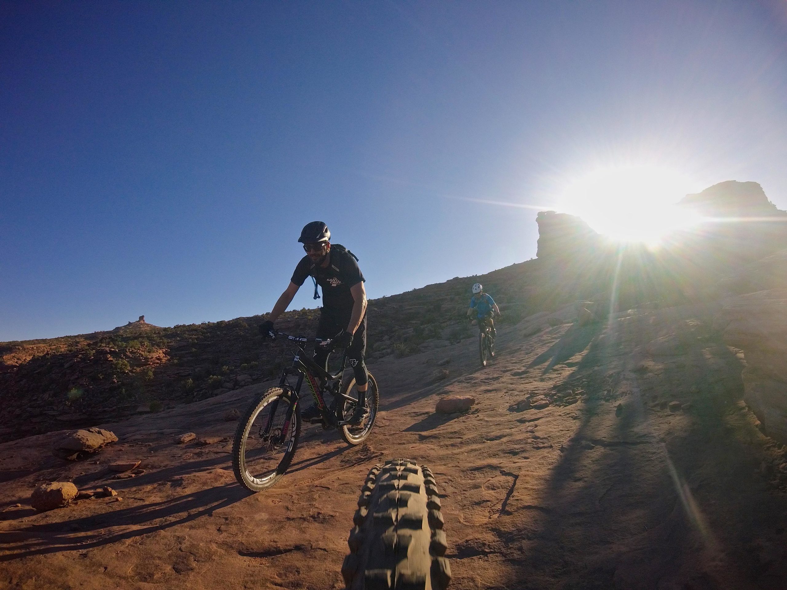 Two mountain bikers navigating a rocky trail under a clear blue sky, with the sun shining brightly in the background. One rider is in a black shirt and helmet, while the other is in a blue shirt. The landscape features red rock formations and scattered vegetation. Captain Ahab mountain bike trail.