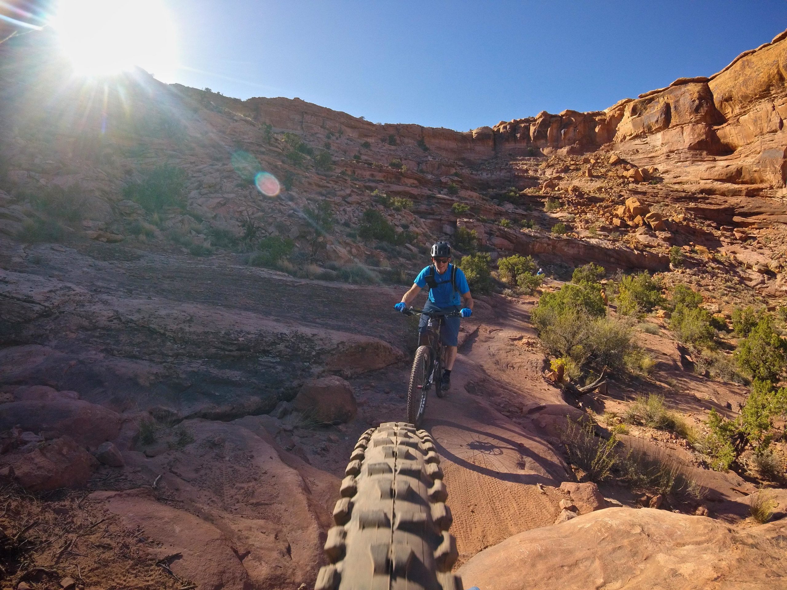 A mountain biker rides along a rocky trail in a sunlit canyon, with the sun shining brightly in the background. The foreground features the wheel of the bike, and the rugged terrain is dotted with shrubs and rocky outcrops. Hymasa mountain bike trail.