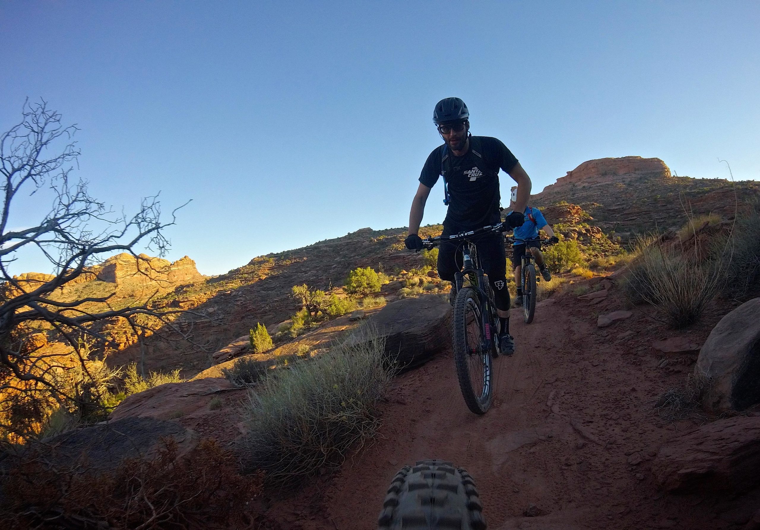 A mountain biker navigates a rocky trail during sunset, with red rock formations and scrub brush surrounding the path. Another cyclist follows closely behind. The sky is clear and blue, indicating a beautiful day for outdoor adventure. Captain Ahab mountain bike trail.