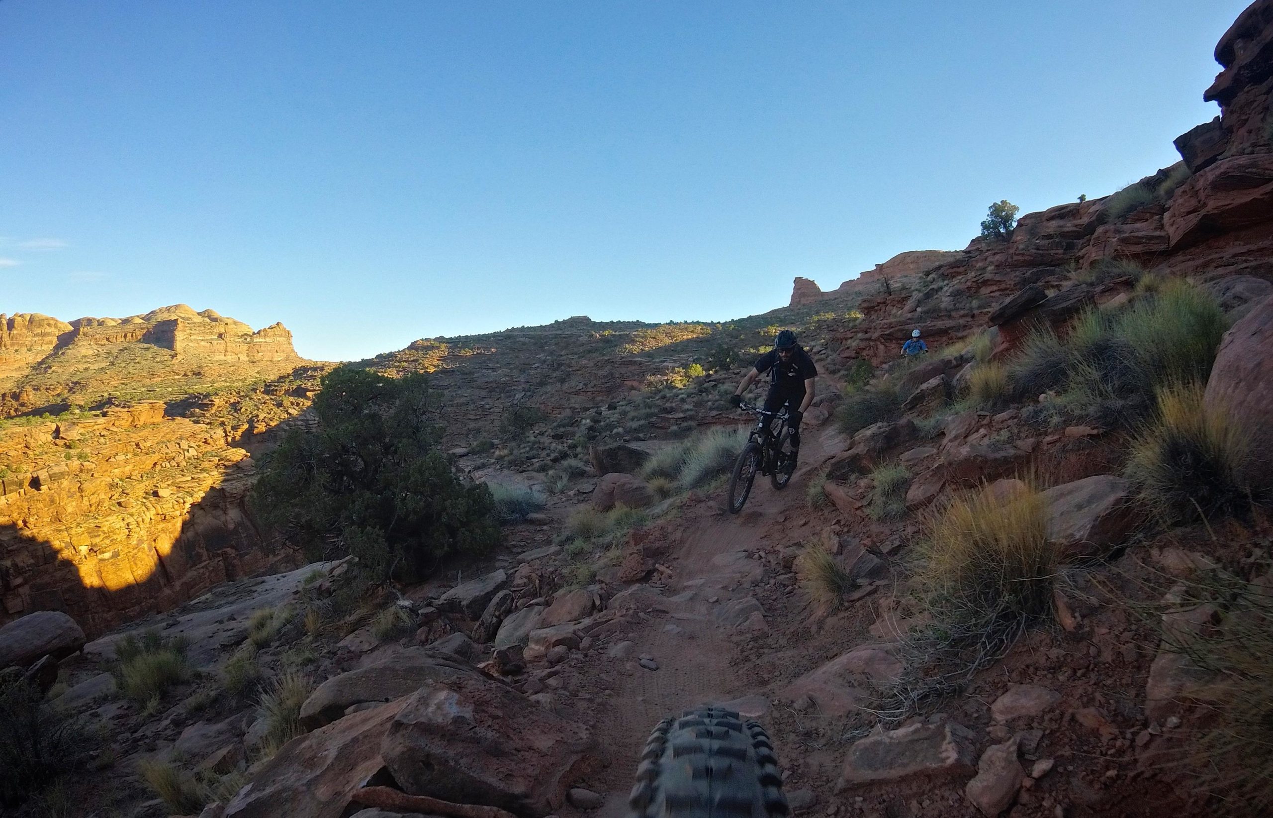 A mountain biker navigating a rocky trail in a desert landscape, surrounded by rugged cliffs and sparse vegetation under a clear blue sky. Captain Ahab mountain bike trail.