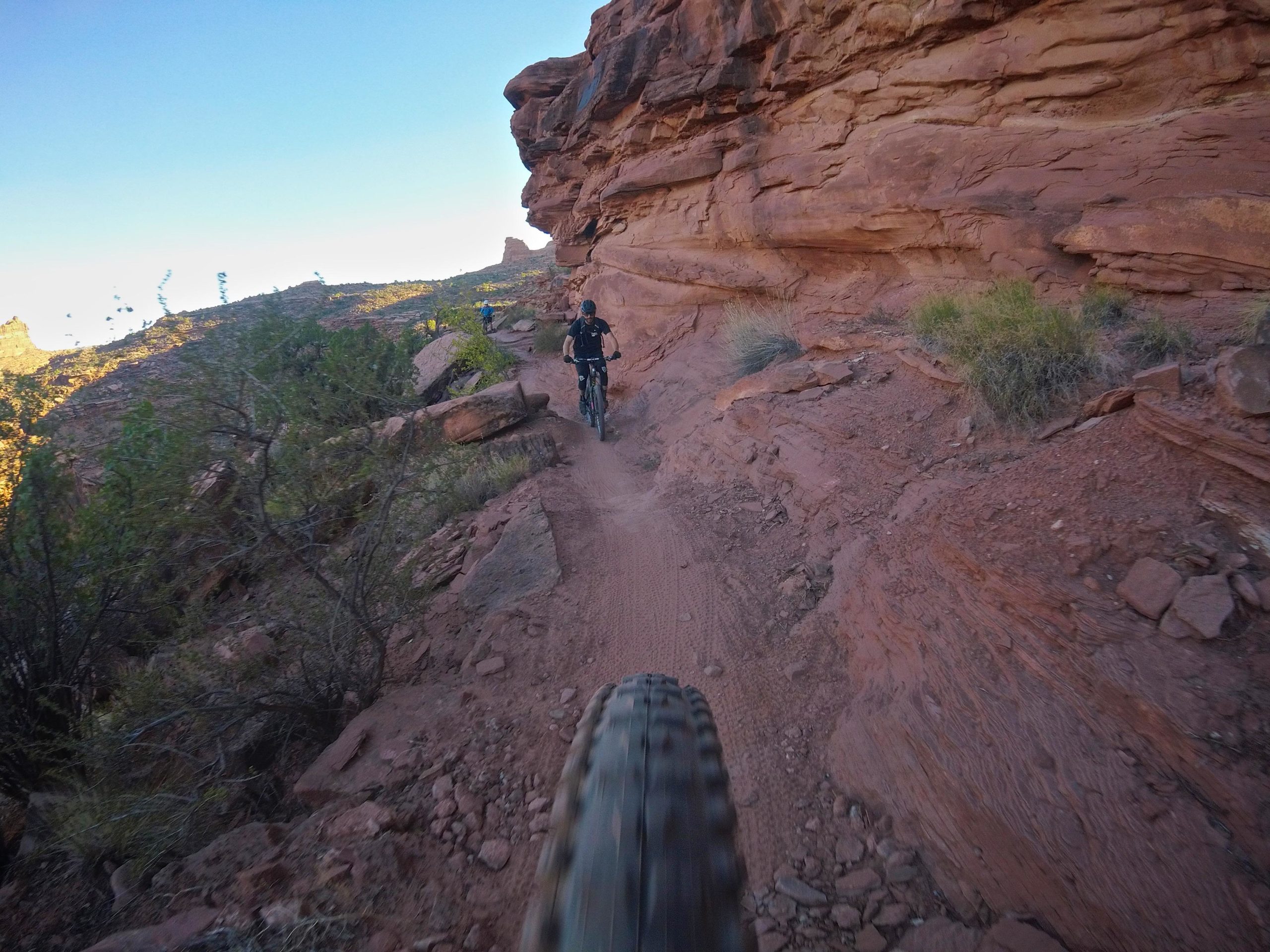 A mountain biker rides along a narrow trail surrounded by rocky terrain and vegetation, with a scenic view of a canyon in the background. The image captures the dynamic motion of biking on a dirt path, emphasizing the rugged landscape and outdoor adventure. Captain Ahab mountain bike trail.