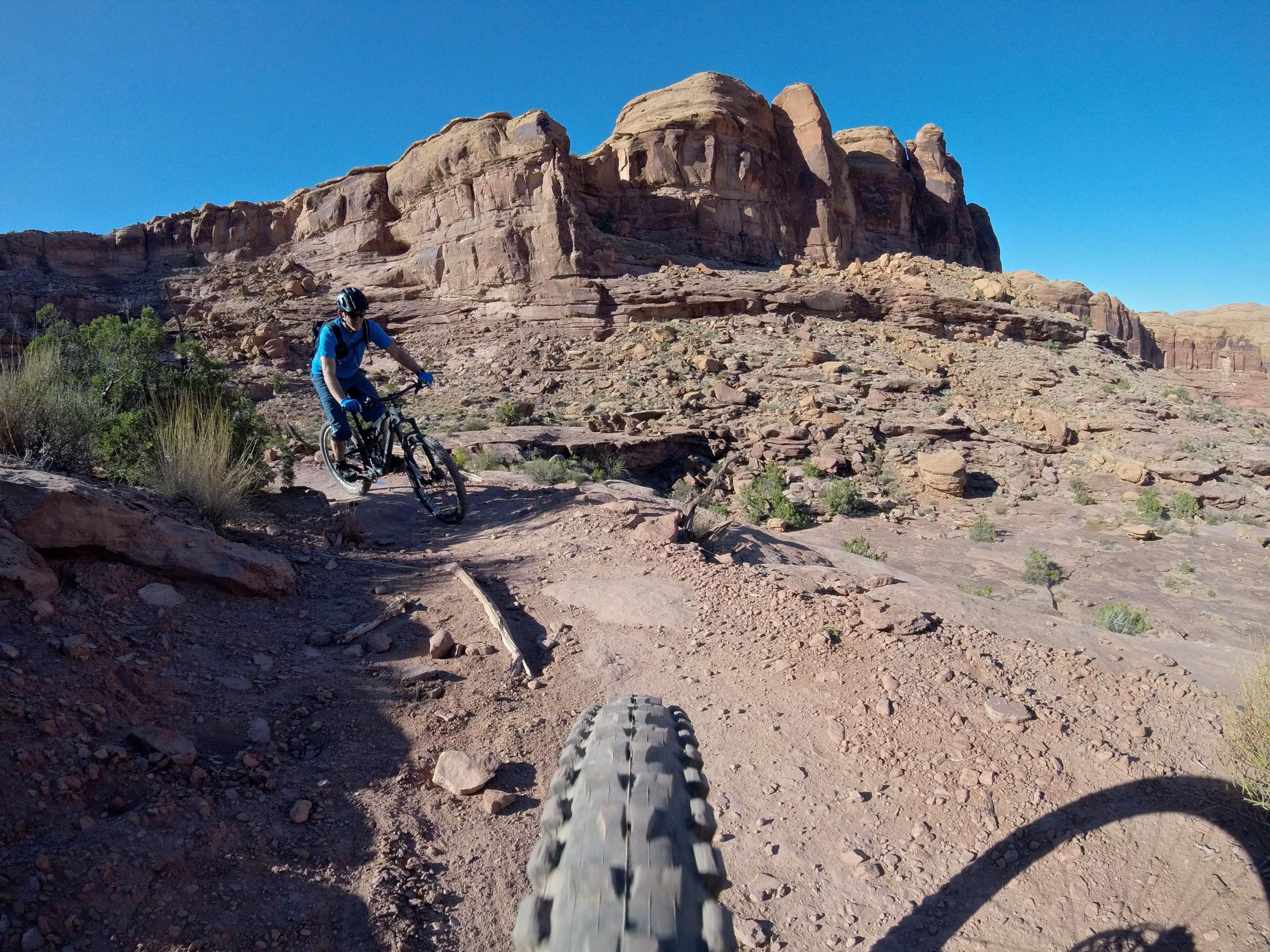 A mountain biker navigating a rocky trail in a desert landscape, with towering red rock formations and a clear blue sky in the background. The close-up view of the bike tire indicates the dynamic action of the ride. Hymasa mountain bike trail.