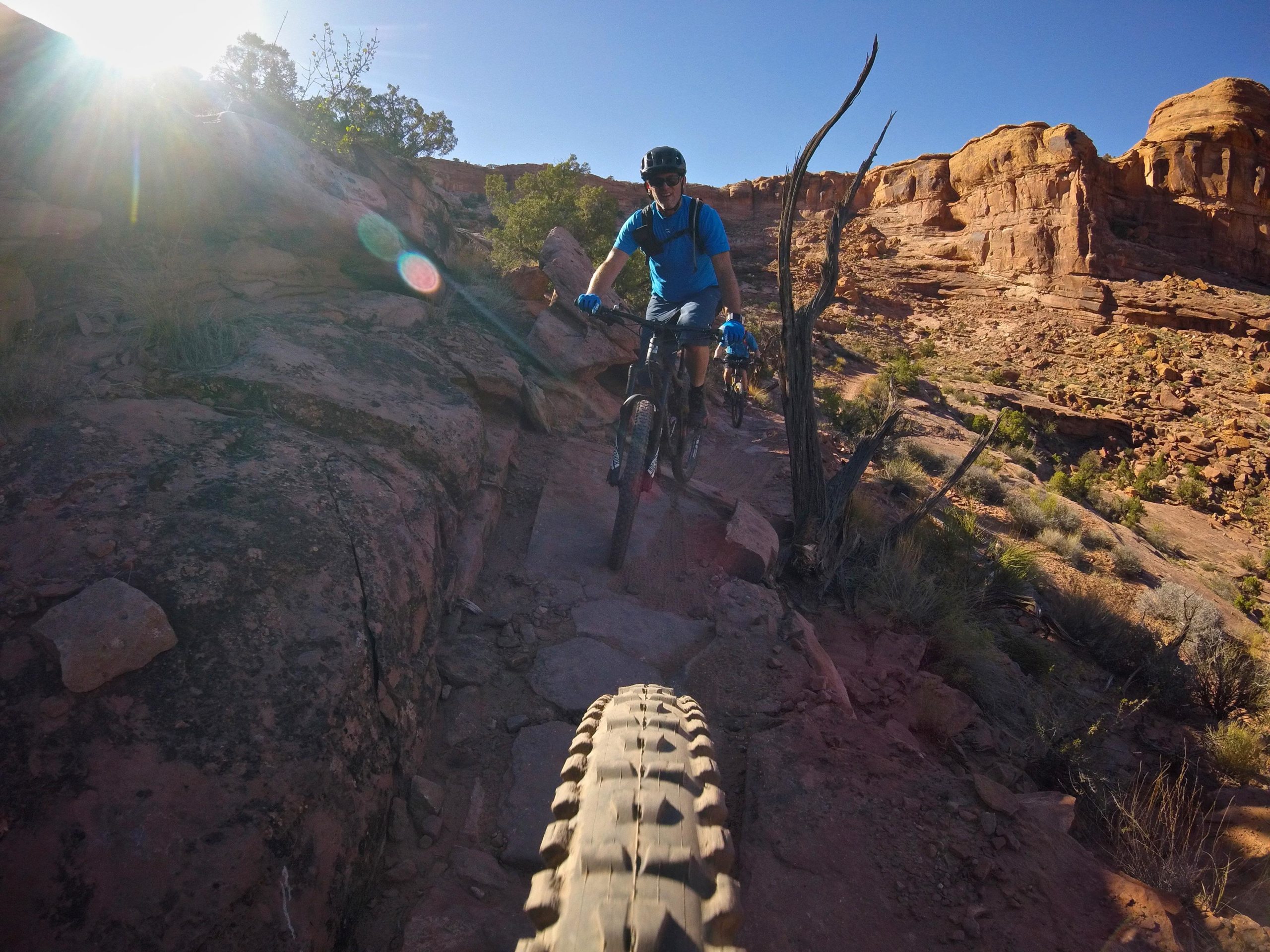A mountain biker rides along a rocky trail in a desert landscape, with sun shining in the background. The foreground features the tire of the bike, while another rider can be seen in the background, making their way through the rugged terrain. The scene captures the beauty of outdoor biking adventures. Hymasa mountain bike trail.