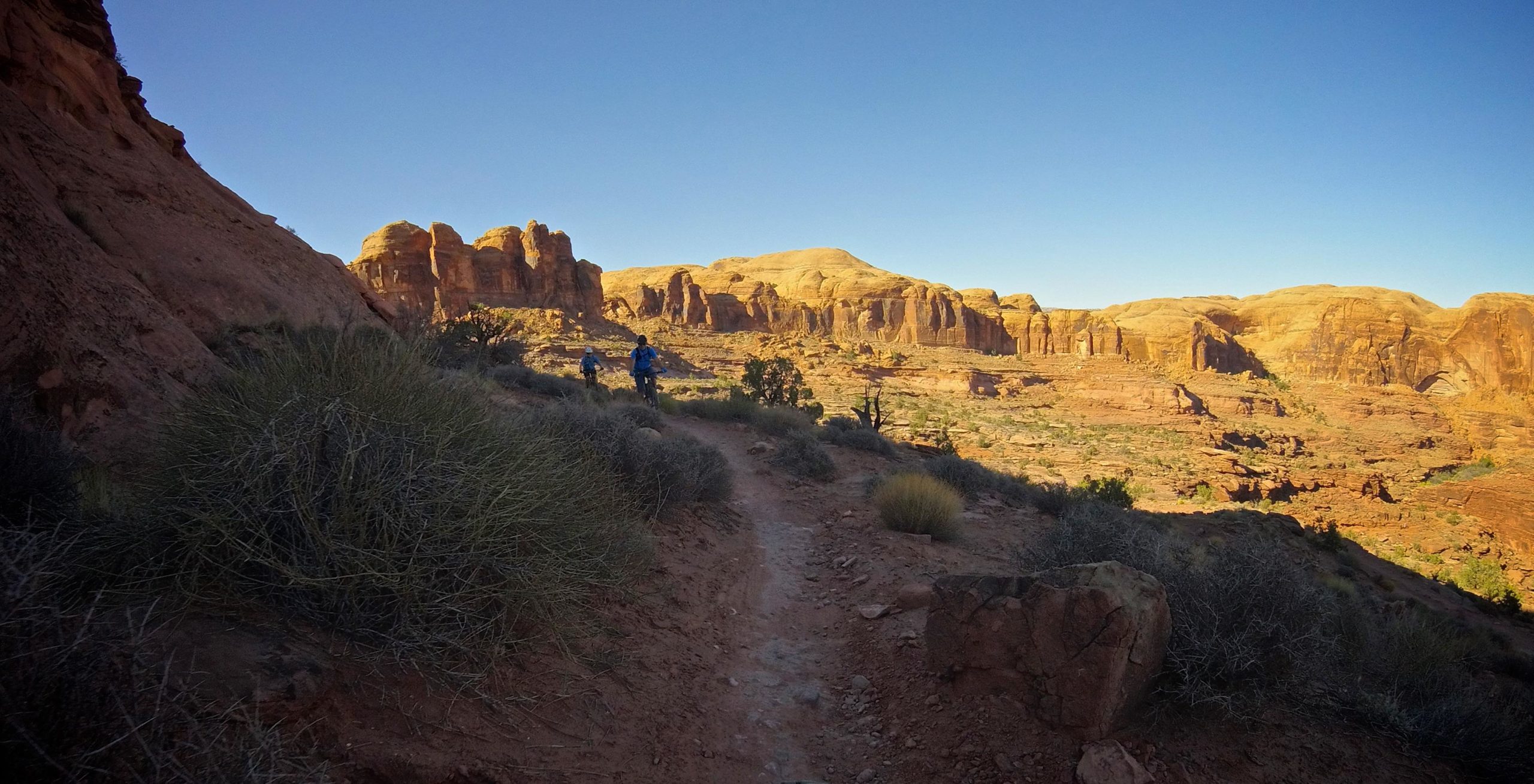 A scenic view of a dirt trail winding through rugged, reddish rock formations under a clear blue sky. Two cyclists in blue shirts ride along the path, surrounded by sparse vegetation and striking rock layers in the background. The landscape conveys a sense of adventure and exploration in a natural setting. Hymasa mountain bike trail.