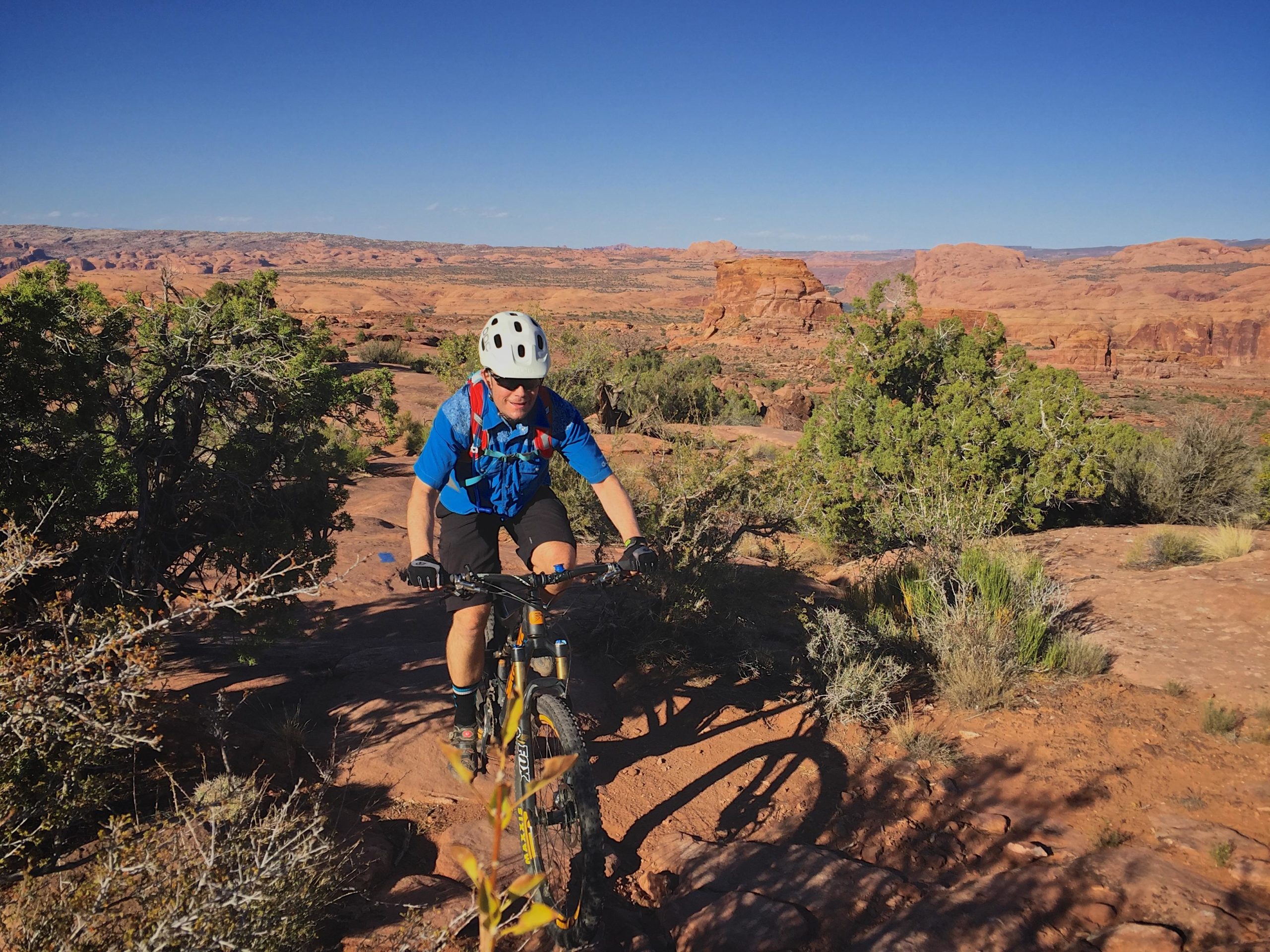 A cyclist wearing a helmet and blue shirt rides a mountain bike along a rocky trail in a rugged desert landscape. The scene features red rock formations and sparse vegetation under a clear blue sky. Captain Ahab mountain bike trail.