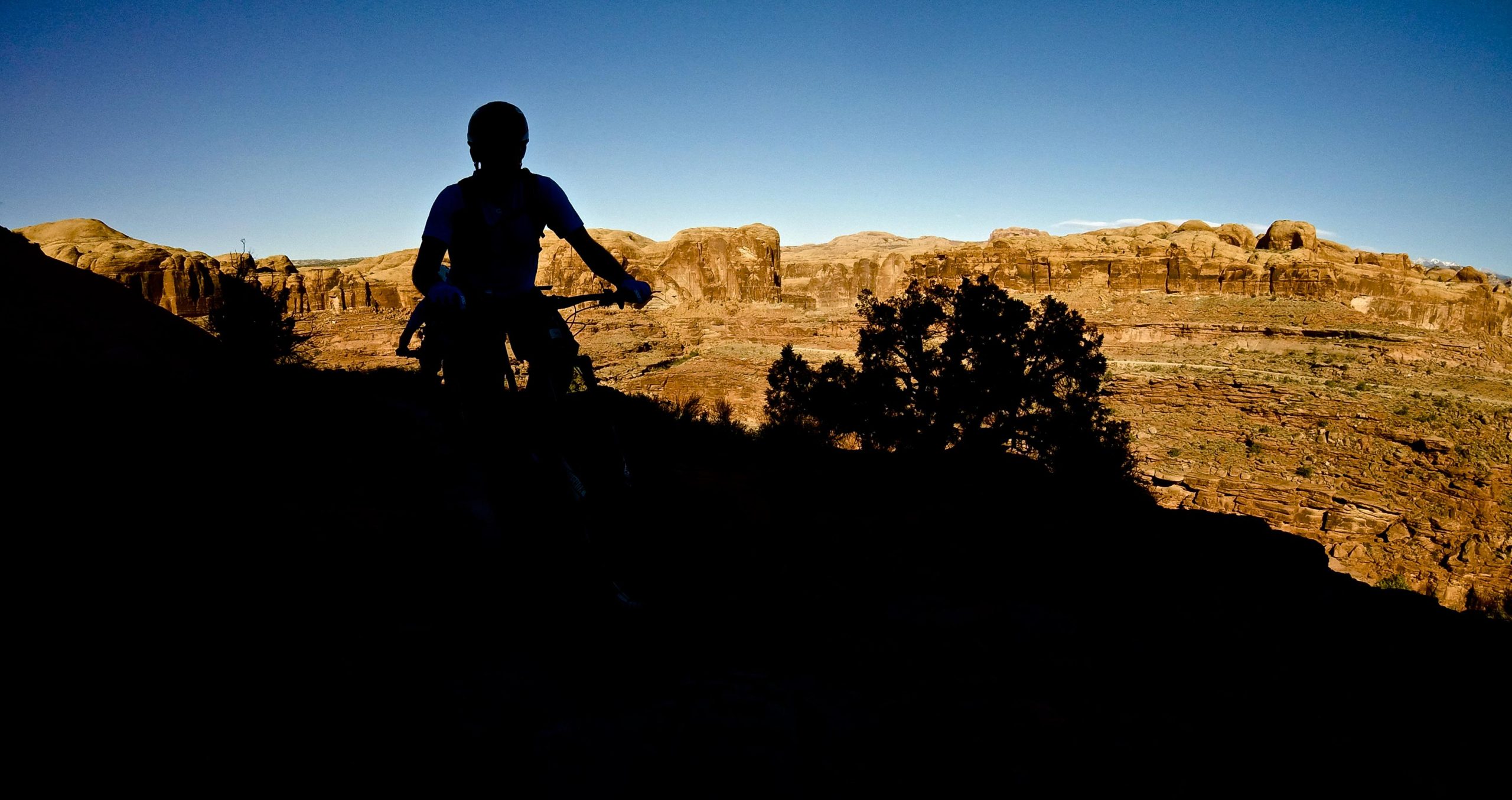 Silhouette of a mountain biker on a rocky trail against a backdrop of rugged cliffs and a clear blue sky during sunset. Hymasa mountain bike trail.