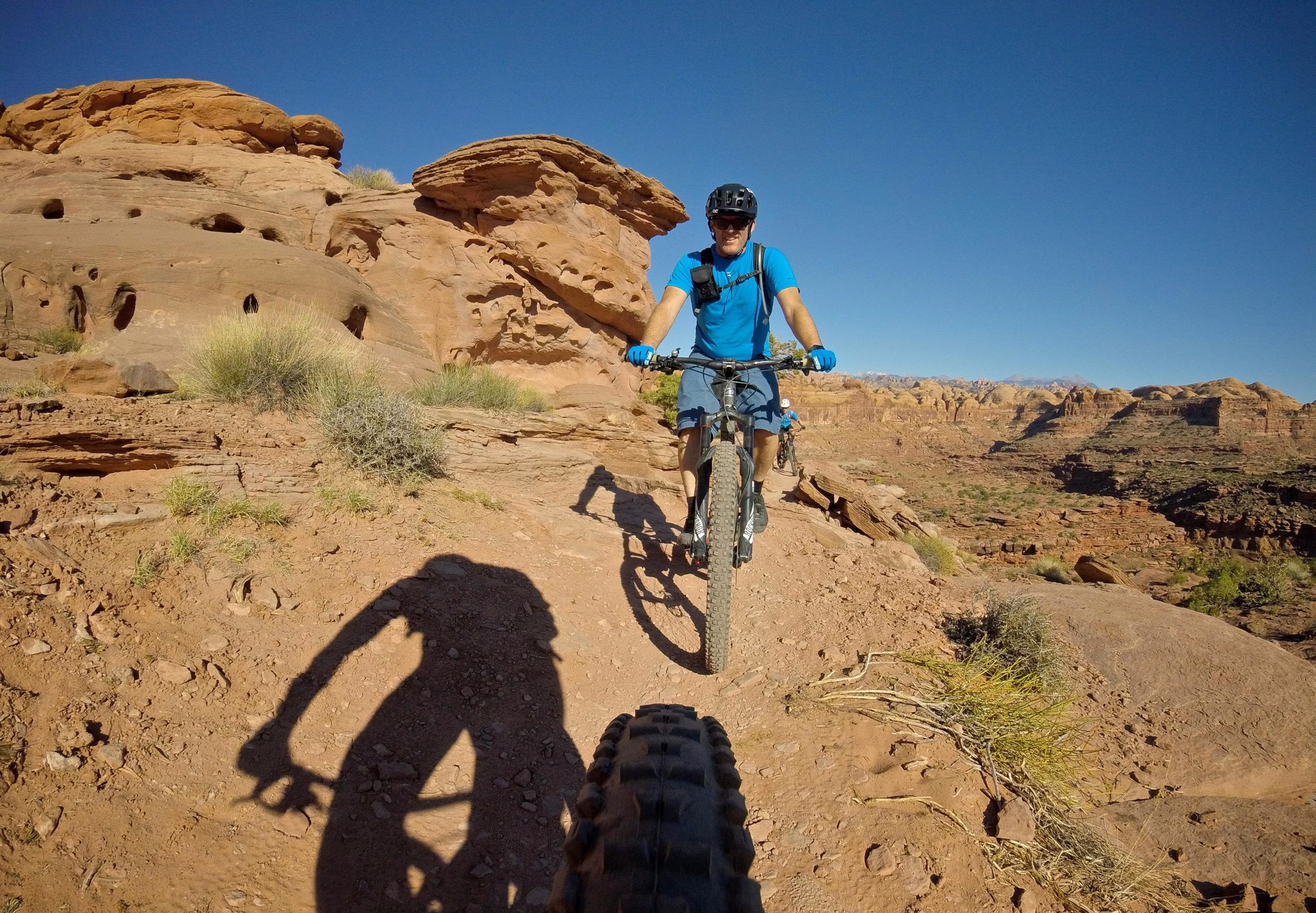 A mountain biker riding along a rocky trail in a desert landscape, with red rock formations and blue sky in the background. The biker is wearing a blue shirt and gloves, and there is a shadow cast on the ground from the bike in the foreground. Hymasa mountain bike trail.
