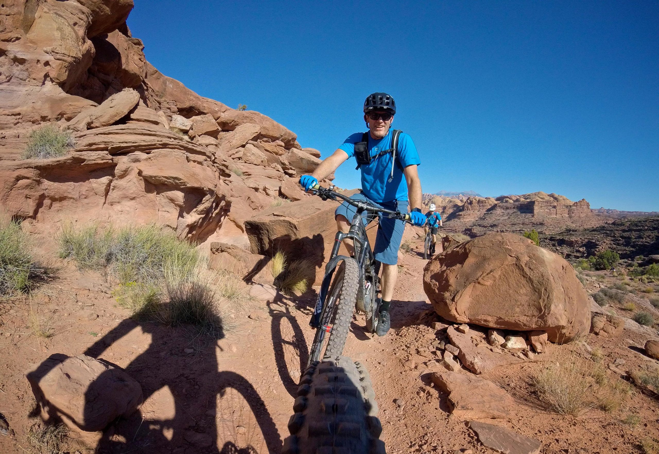 A mountain biker in a blue shirt and gloves rides on a rocky trail, surrounded by red rock formations and a clear blue sky. In the background, another biker is seen navigating the terrain. Hymasa mountain bike trail.