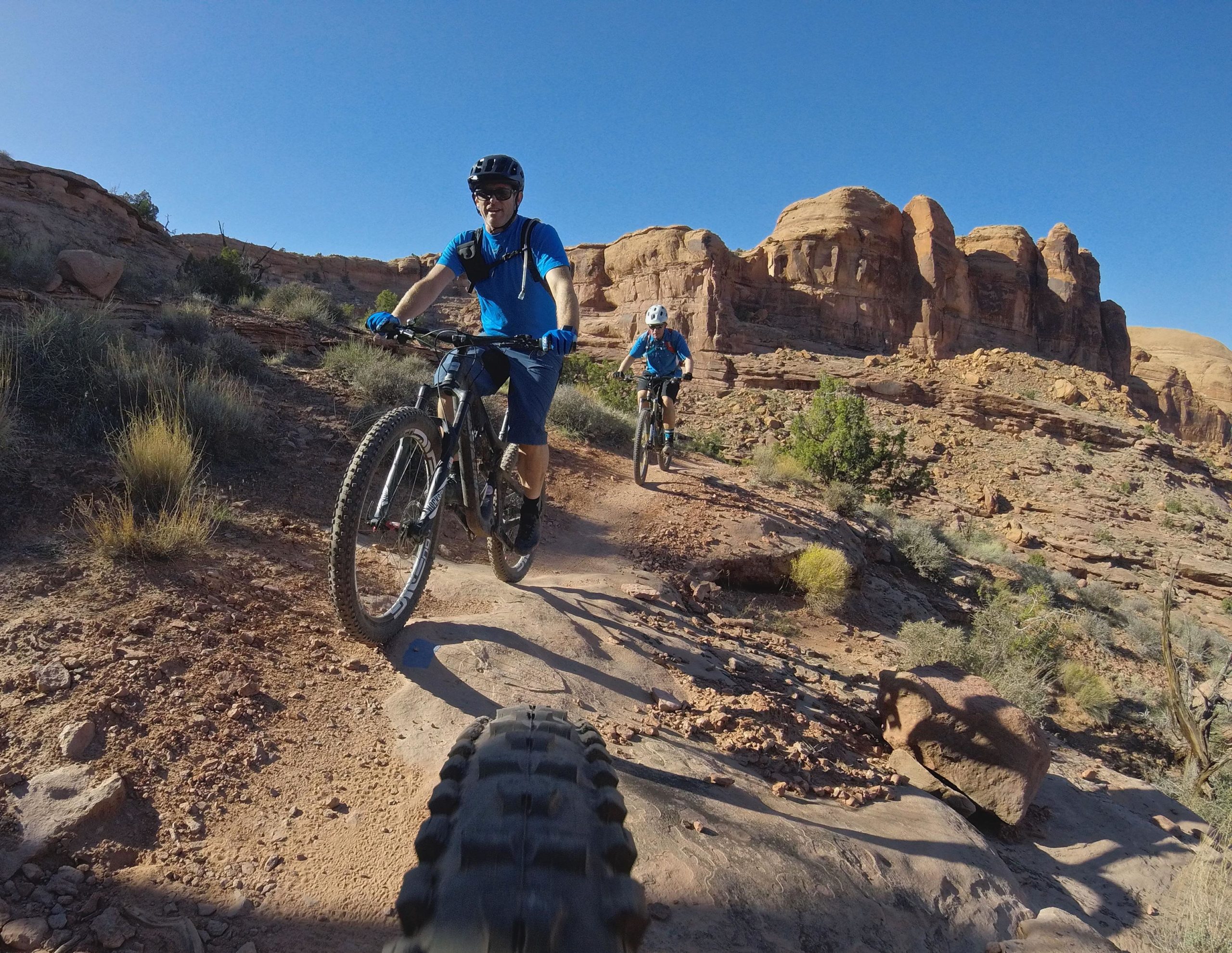 Two mountain bikers ride along a dirt trail surrounded by rugged rock formations and sparse vegetation. One biker, wearing a blue shirt and helmet, is in the foreground, with a close-up view of a bike tire visible in the lower part of the image. The other biker trails behind, also dressed in blue. The scene is set against a clear blue sky, showcasing a sunny day outdoors. Hymasa mountain bike trail.