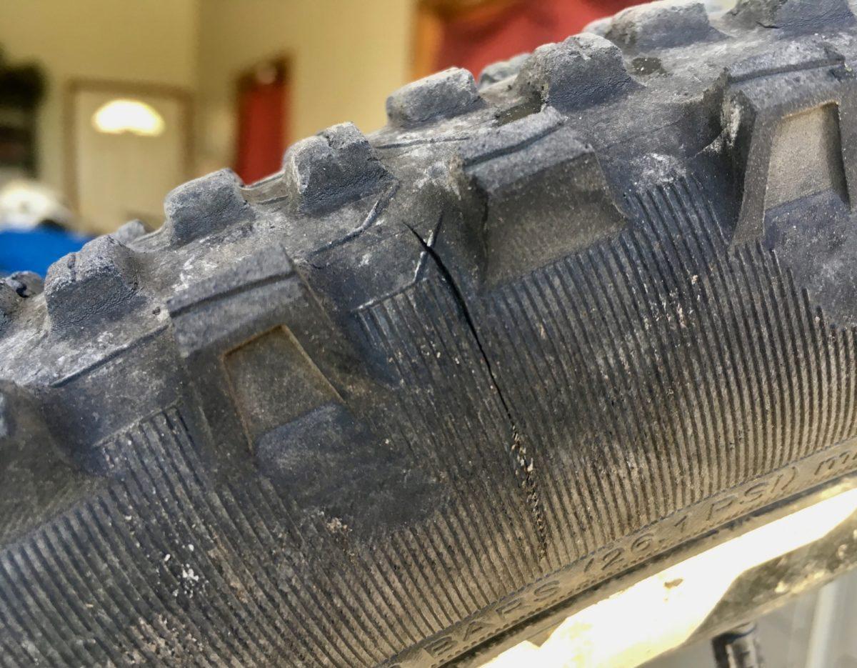 Michelin Wild AM: Close-up of a damaged bicycle tire showing a noticeable crack in the tread. The tire features prominent treads and markings, indicating wear and use. The background is blurred, suggesting an indoor setting.