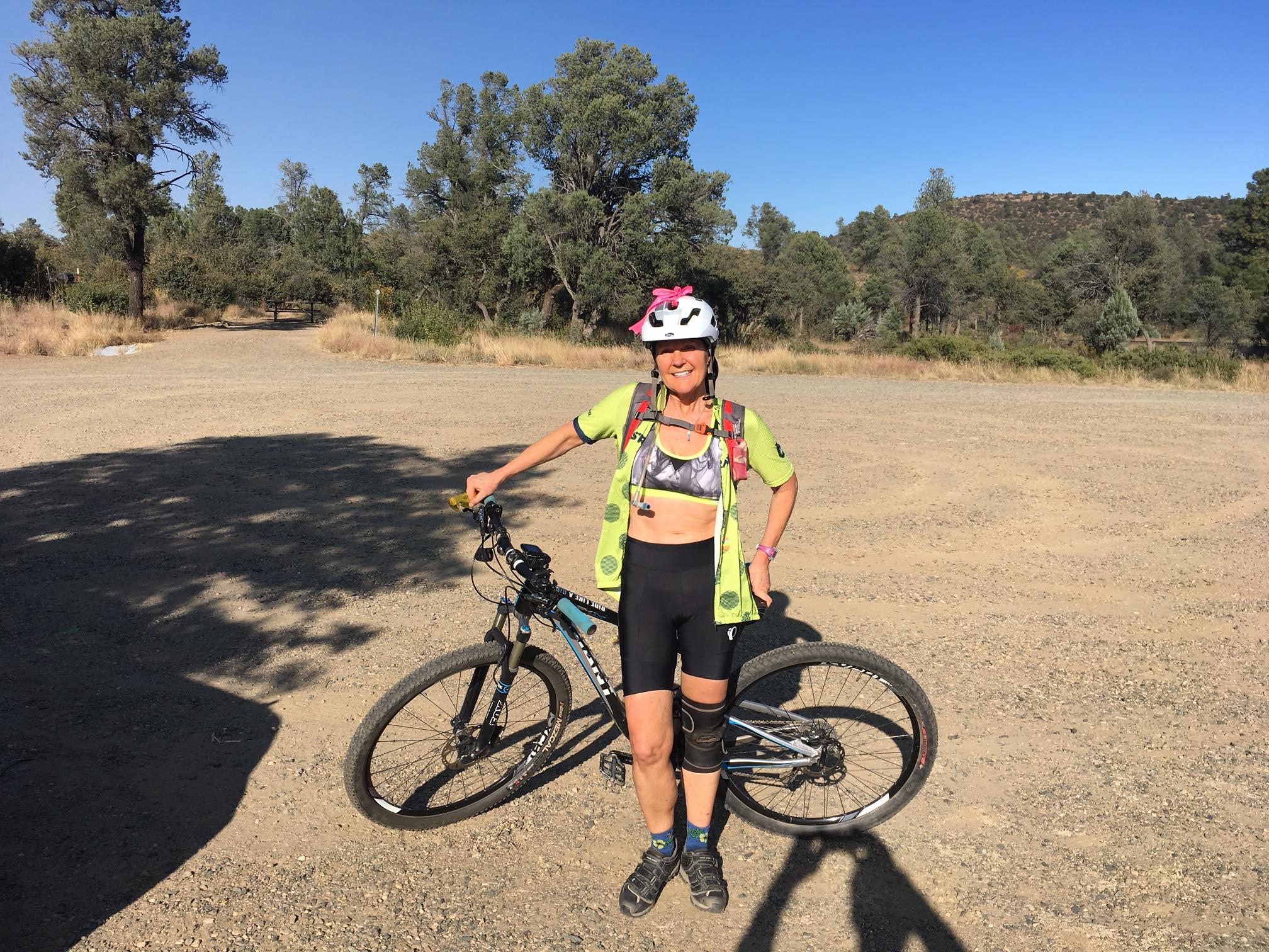 A woman wearing a helmet and athletic gear stands next to her mountain bike on a gravel pathway surrounded by trees. She is smiling, dressed in a sports top and shorts, and has a backpack on her back. The background features sparse vegetation and a clear blue sky. Ranch Trail 62 mountain bike trail.